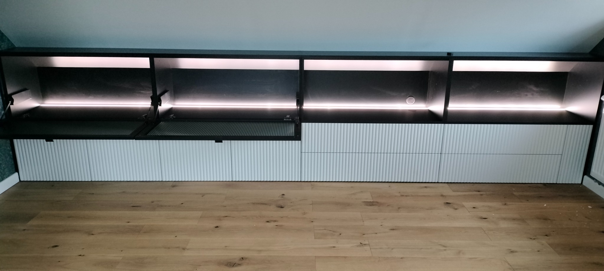 Szafa wnękowa na poddaszu z oświetleniem LED, białe fronty z frezowaniem pionowym, czarne korpusy górne, otwarty schowek z kratką wentylacyjną.