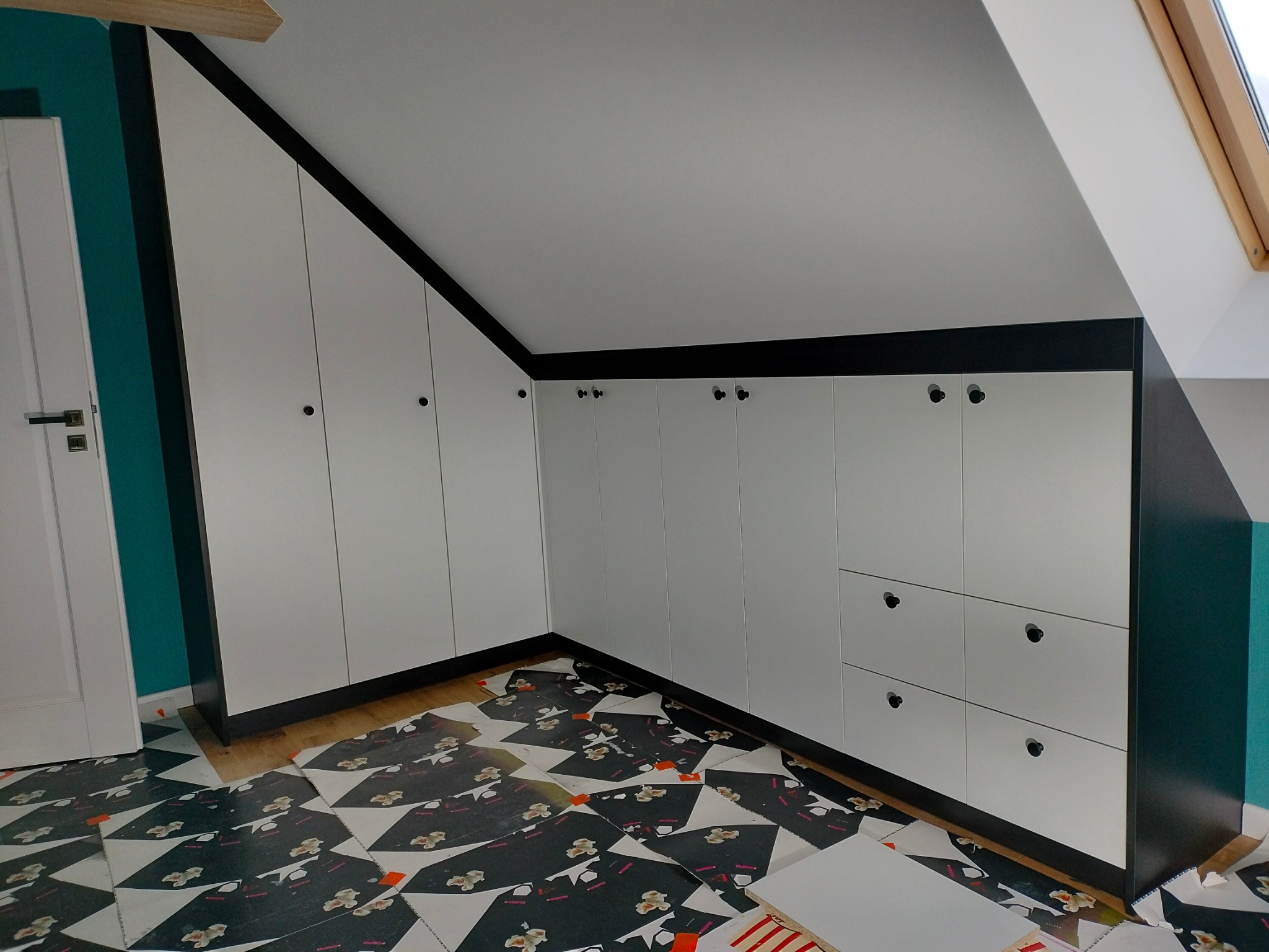 Biała szafa wnękowa z czarnymi obramowaniami i okrągłymi uchwytami, dopasowana do skosu dachu. Na podłodze zabezpieczenie z kartonu.