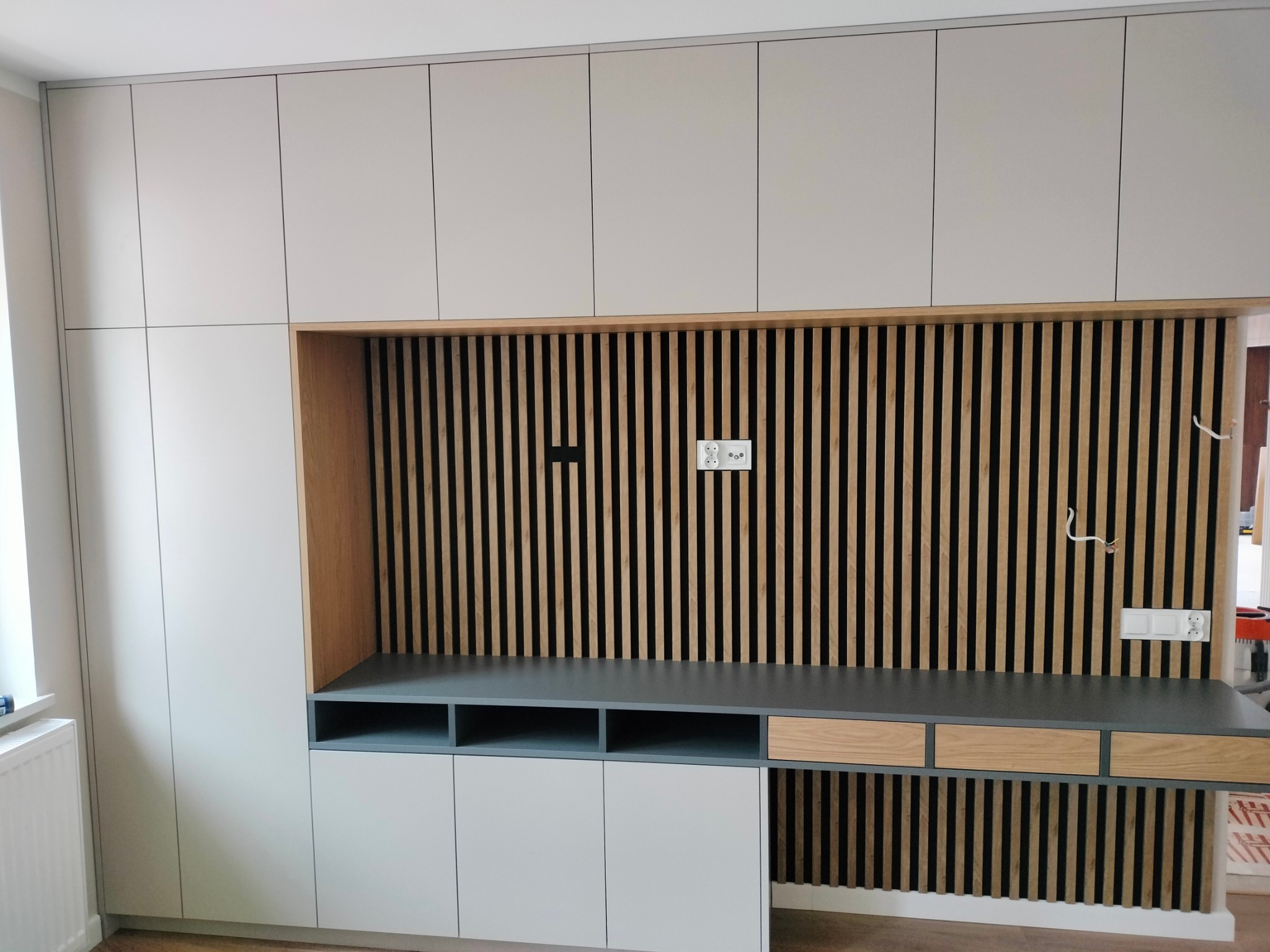 Jasnoszara zabudowa meblowa z drewnianymi lamelami na ścianie, szary blat i białe szafki dolne. Widoczne gniazdka elektryczne i włączniki.
