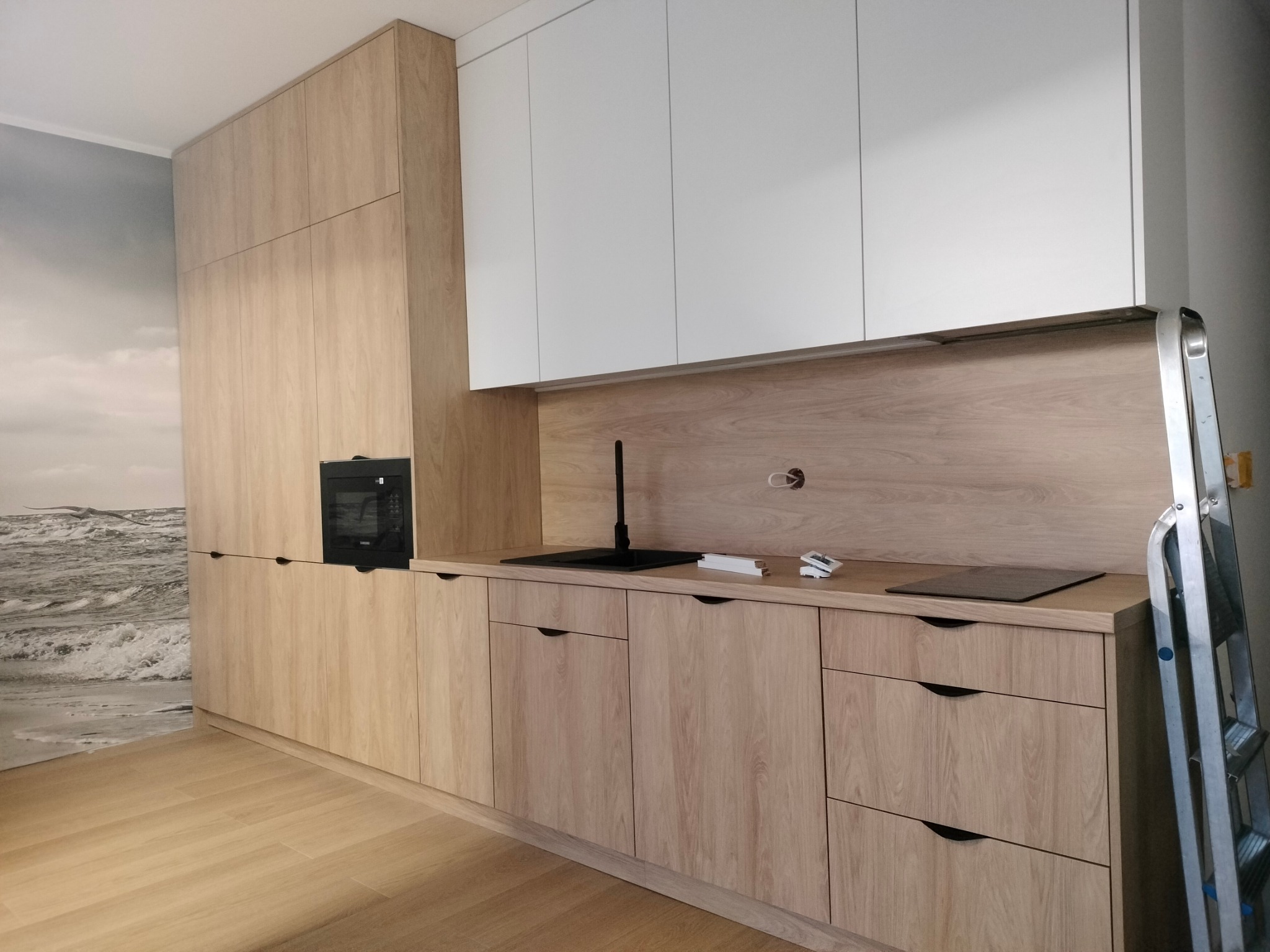 Jasna, nowoczesna kuchnia z drewnianymi frontami i białymi szafkami wiszącymi. Widoczna drabina sugeruje trwający montaż. Na ścianie fototapeta z motywem morza.