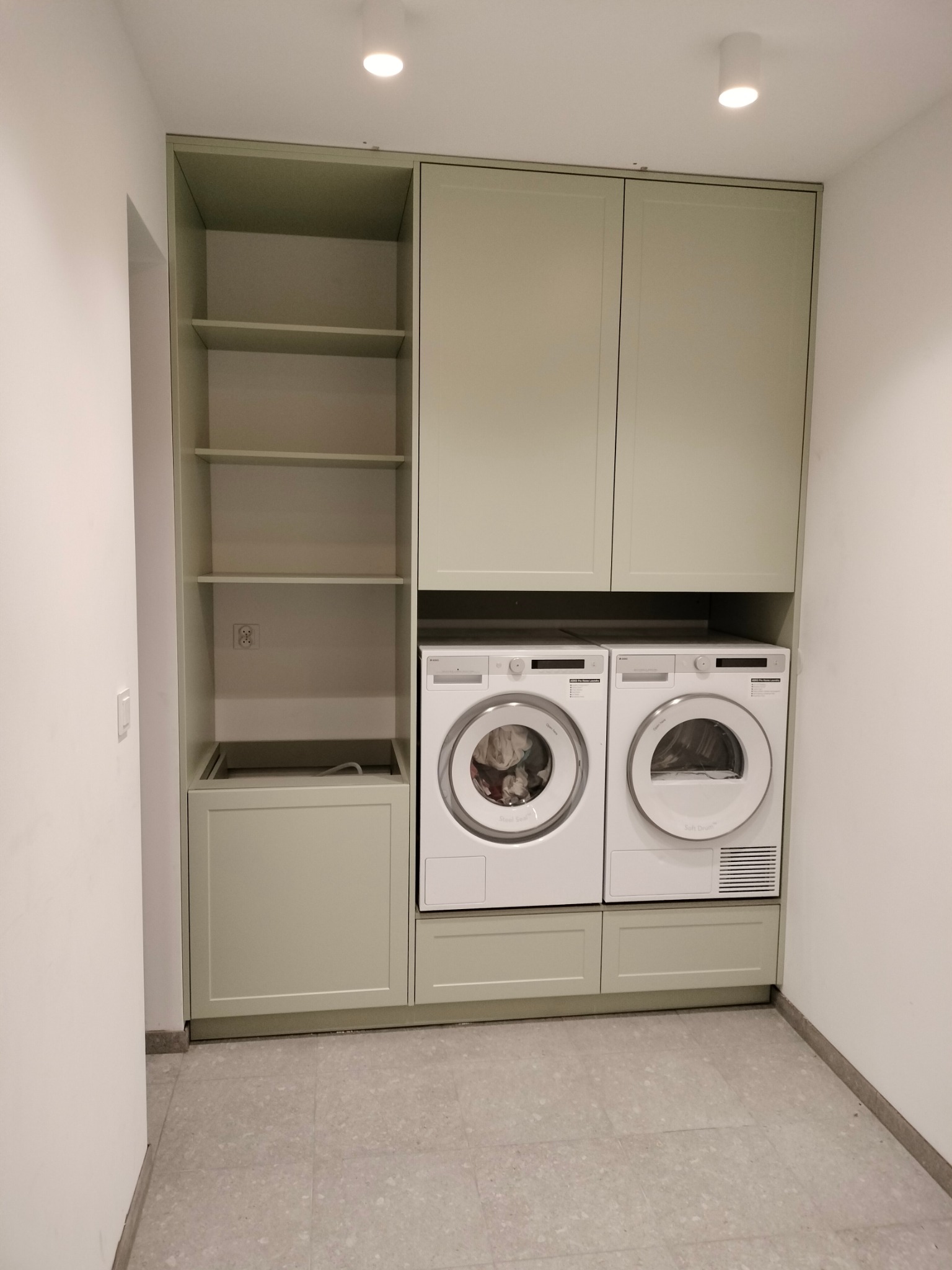 Jasnozielona zabudowa meblowa z szafkami nad pralką i suszarką. Po lewej regał z półkami. Minimalistyczny styl, neutralne tło. Oświetlenie sufitowe.