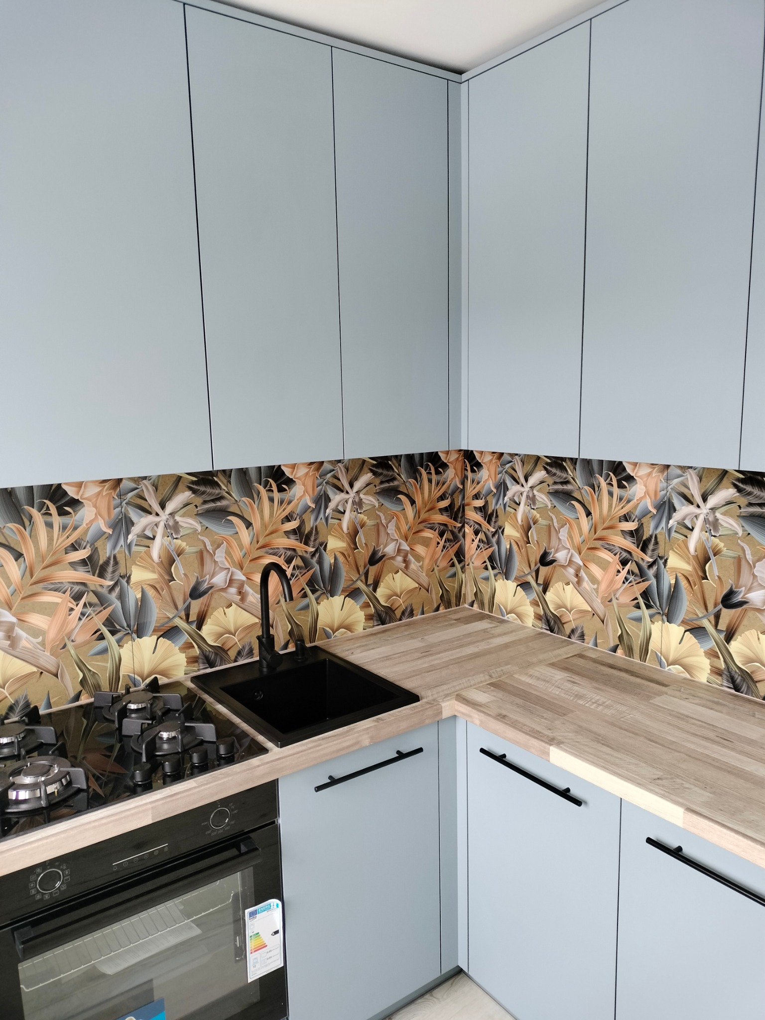 Narożna kuchnia z jasnoniebieskimi frontami, drewnianym blatem i panelem ściennym w motywy roślinne. Czarny zlew i płyta grzewcza. Minimalistyczne, podłużne uchwyty.