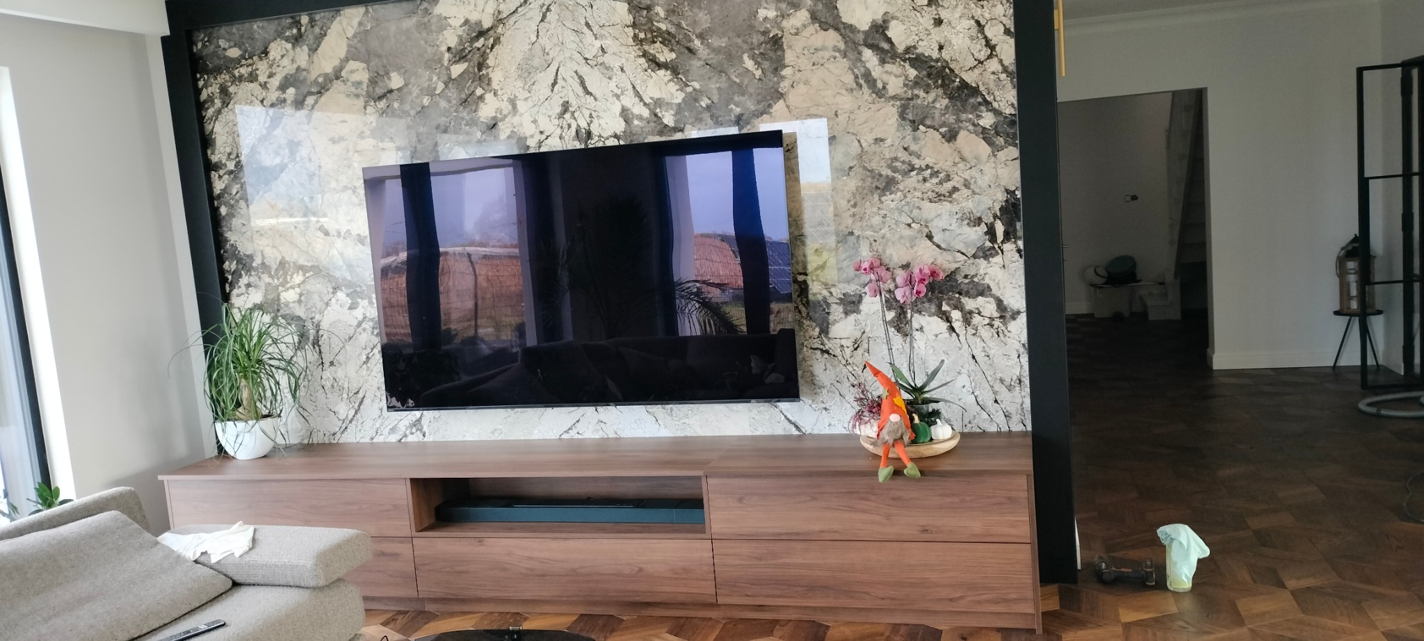 Nowoczesny salon z szafką RTV z drewna orzechowego pod telewizorem, na tle ściany z marmurowym wzorem. Wnętrze dopełniają rośliny i dekoracje.