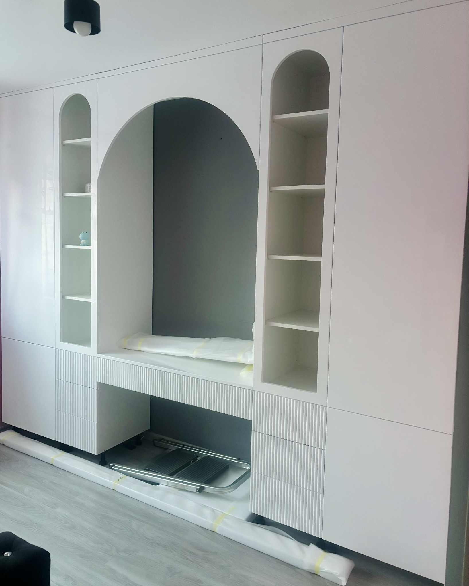 Biała zabudowa meblowa z biurkiem i półkami w kształcie łuków, widoczne zabezpieczenia transportowe, nowoczesny design z frezowanymi frontami.