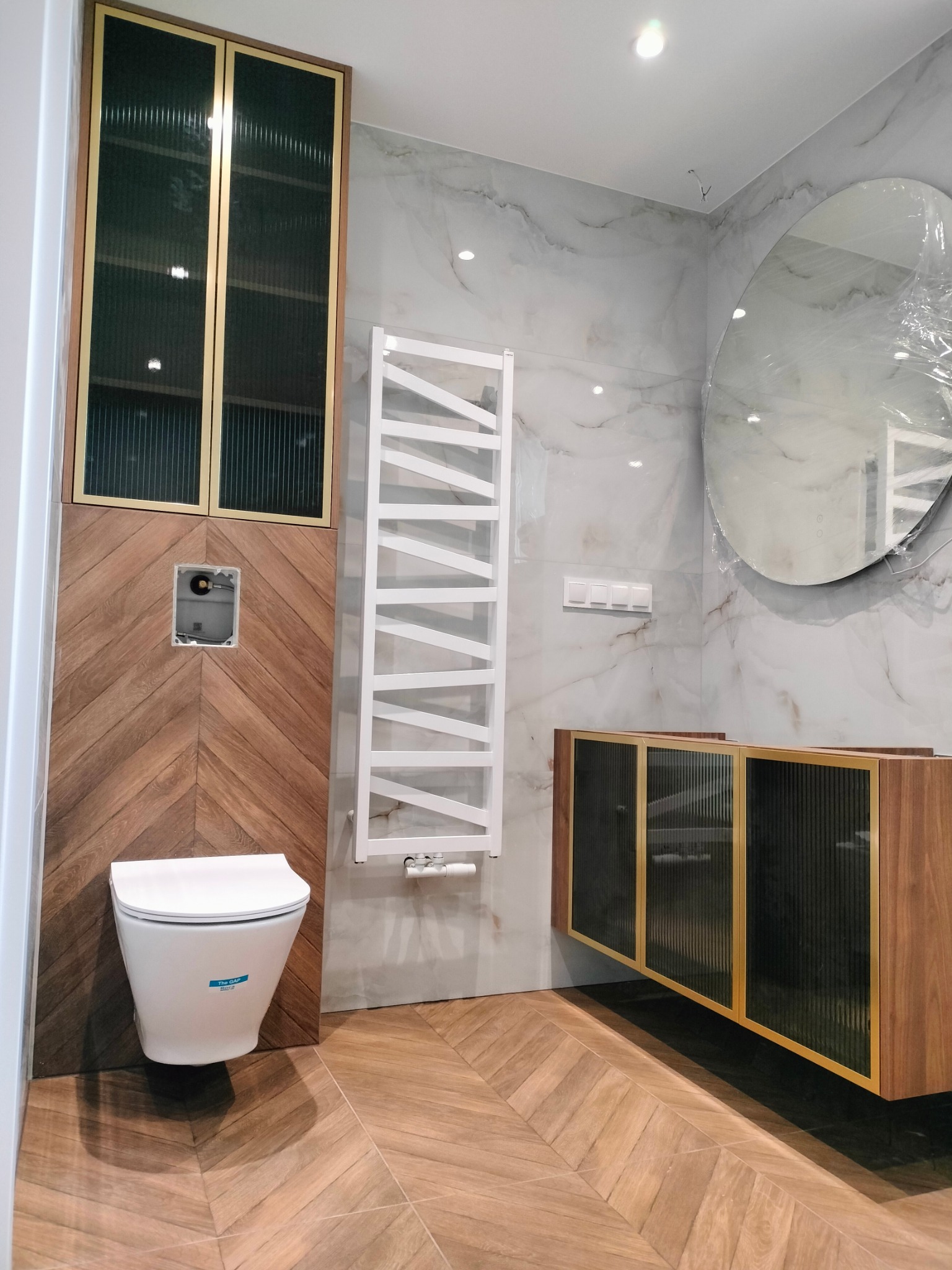 Nowoczesna łazienka z drewnianymi akcentami: szafka pod umywalkę i szafka wisząca z ryflowanymi szybami, biały grzejnik drabinkowy i okrągłe lustro w folii.