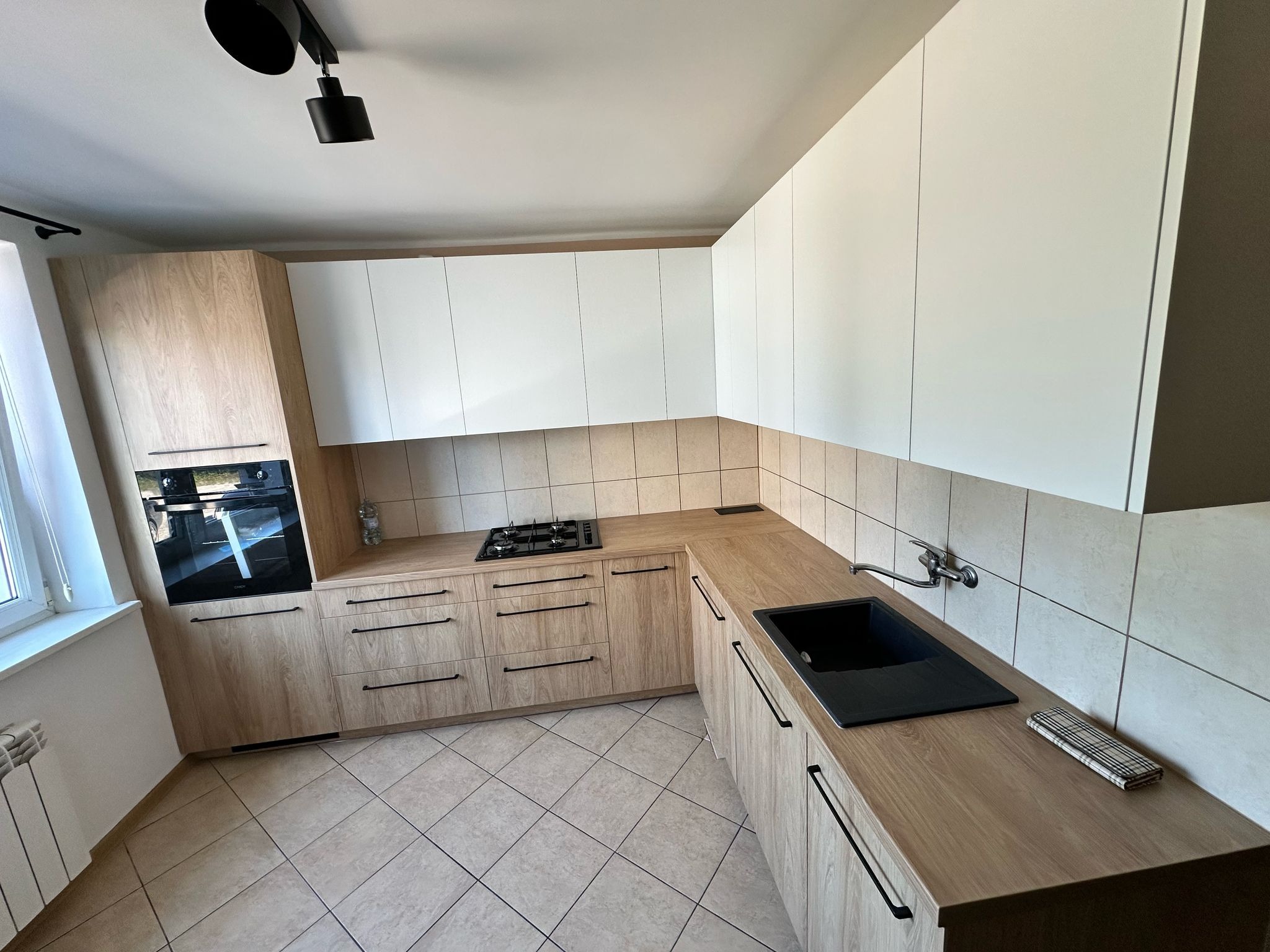 Jasna kuchnia z drewnianymi i białymi szafkami, czarnym zlewem i płytą gazową. Realizacja w Połczynie-Zdroju. Uwagę przykuwają długie, czarne uchwyty.