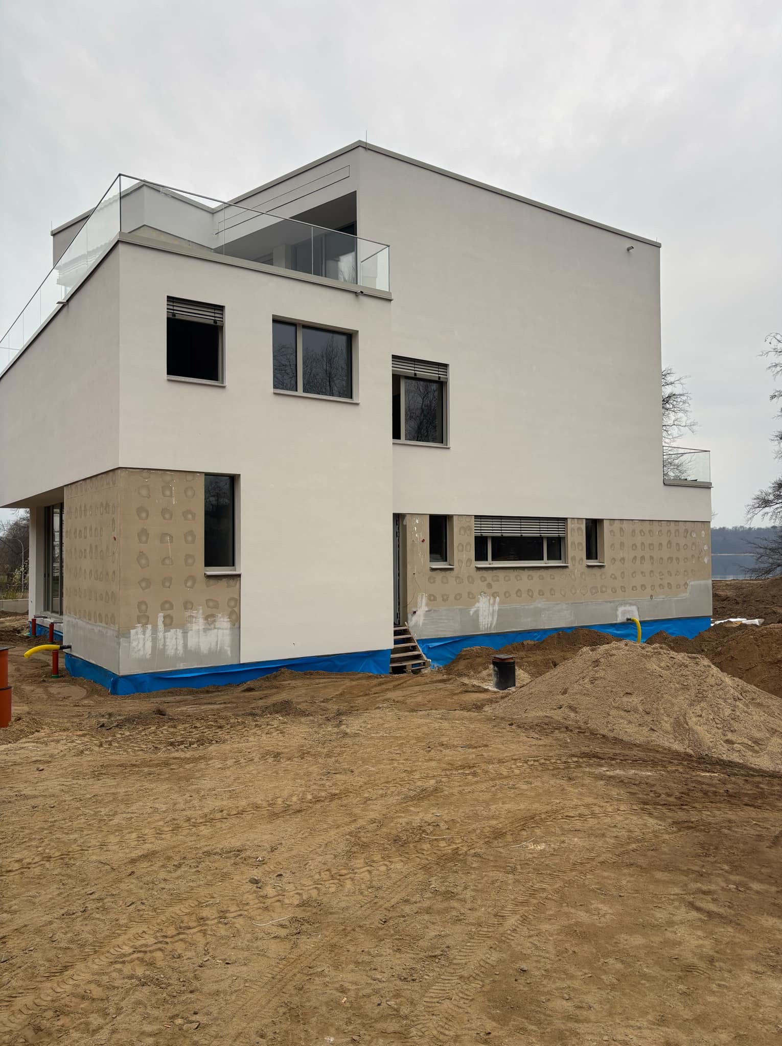 Nowoczesny dom w trakcie prac. Część elewacji ocieplona z widocznymi otworami, reszta pomalowana na biało. Widoczne zabezpieczenia i piasek. Szklane balustrady.