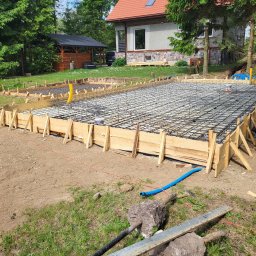 Dawid - Drewniany szalunek zbrojony stalową siatką i rurami instalacyjnymi, przygotowany pod wylewanie fundamentu, na tle domu z czerwonym dachem.