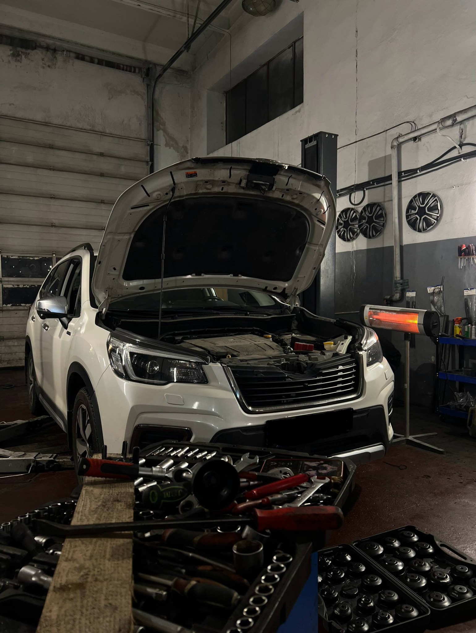 Biały samochód z otwartą maską w warsztacie. Widoczne narzędzia, podnośnik i lampa grzewcza. Diagnostyka silnika w trakcie.