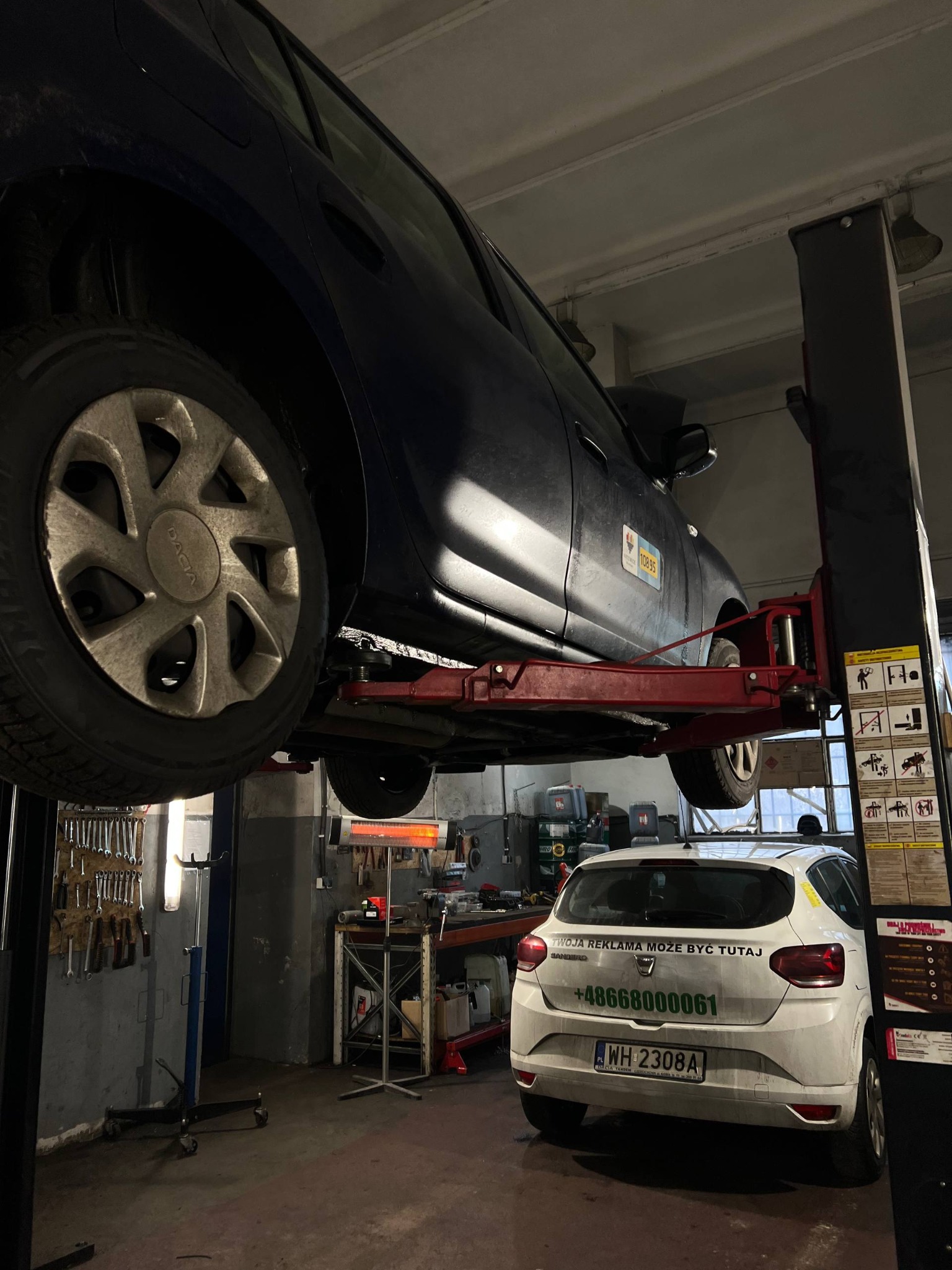Samochód Dacia na podnośniku w warsztacie, widoczny drugi pojazd z reklamą na tylnej klapie. W tle narzędzia i wyposażenie warsztatowe.