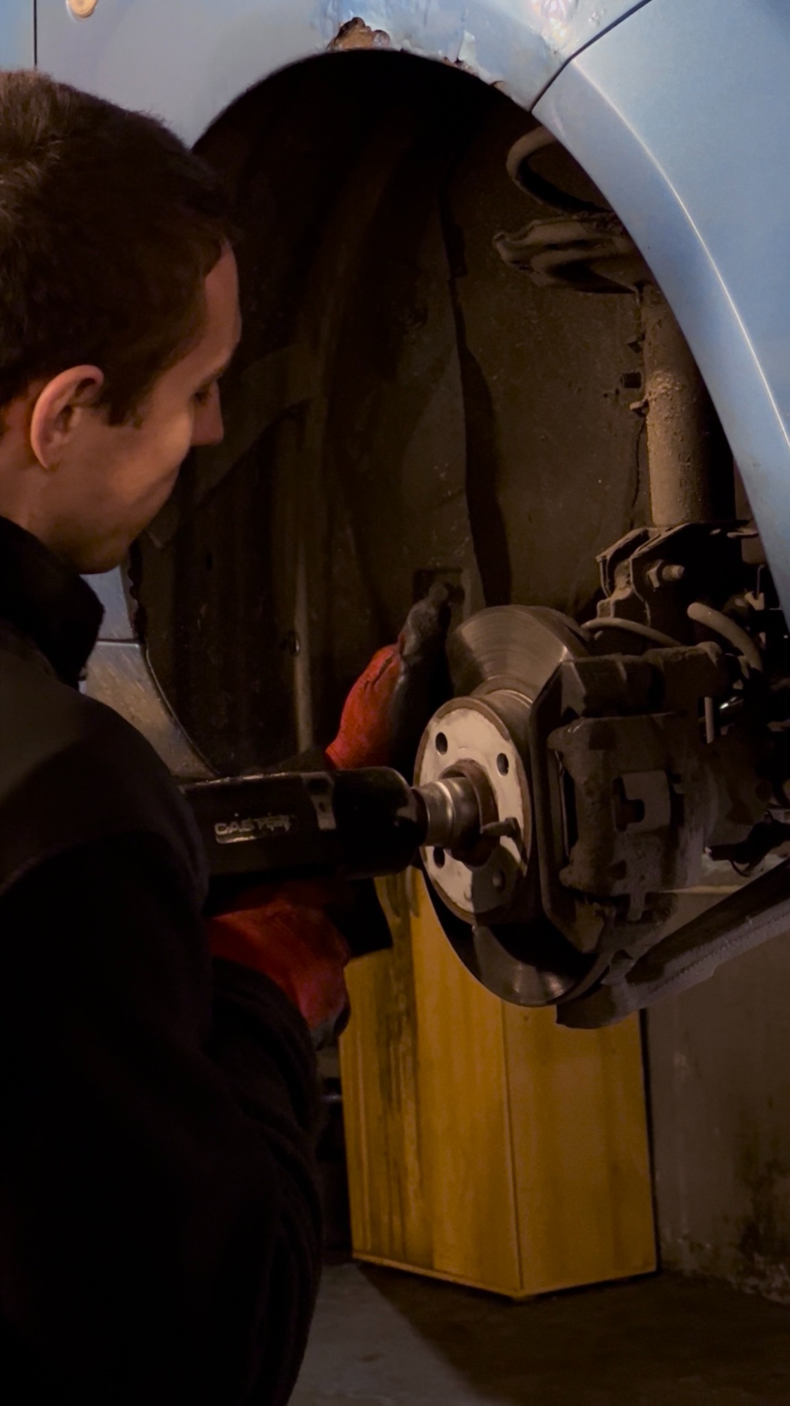 Mechanik w czerwonych rękawicach serwisuje układ hamulcowy samochodu, używając narzędzia. Widoczny element zawieszenia i fragment karoserii.