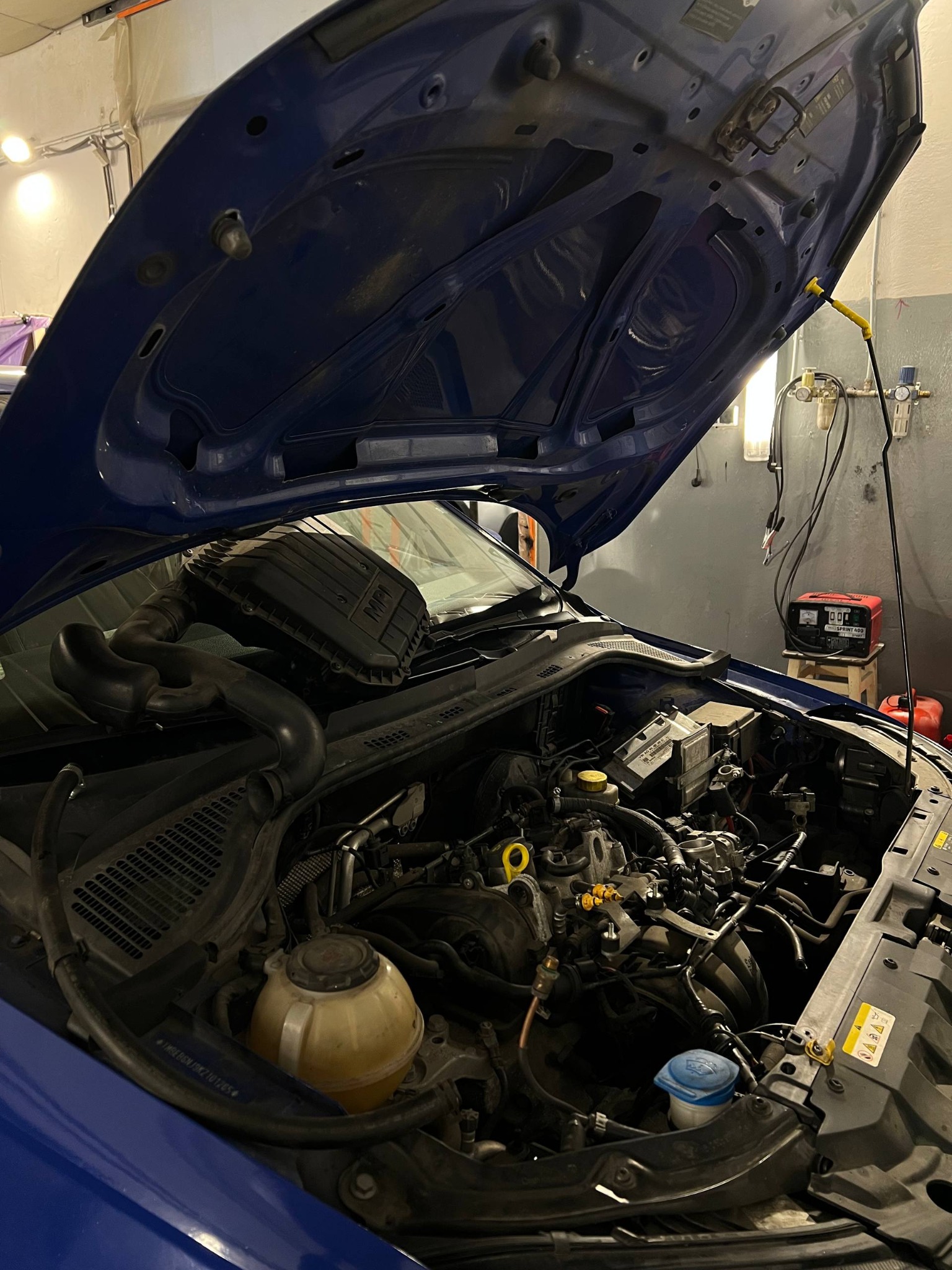 Otwarta maska niebieskiego samochodu w warsztacie. Widoczny silnik, akumulator, przewody i inne elementy. Diagnostyka lub naprawa w toku.