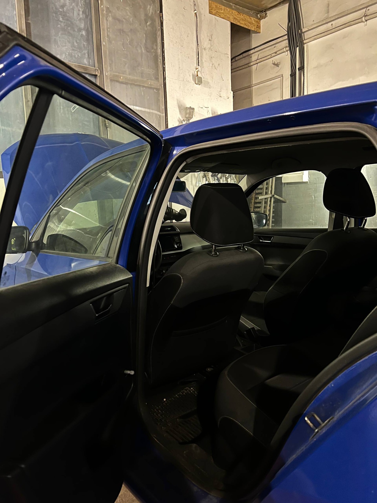Wnętrze niebieskiego samochodu z otwartymi drzwiami, widoczne fotele i deska rozdzielcza. Otwarta maska sugeruje serwis lub naprawę.
