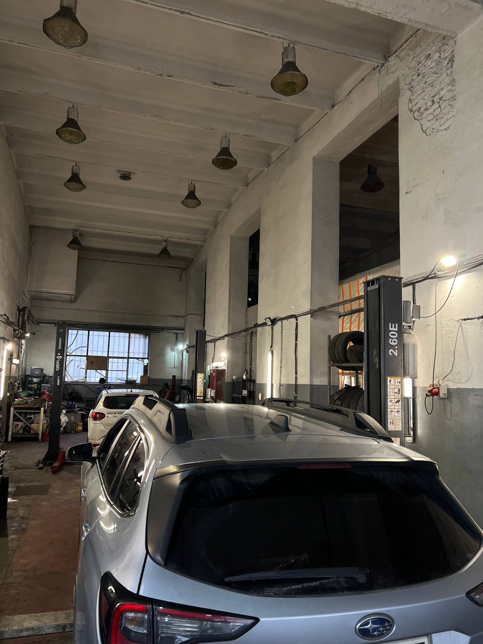 Wnętrze warsztatu samochodowego z podnośnikami i srebrnym Subaru na pierwszym planie. Widoczne lampy przemysłowe i surowe ściany.
