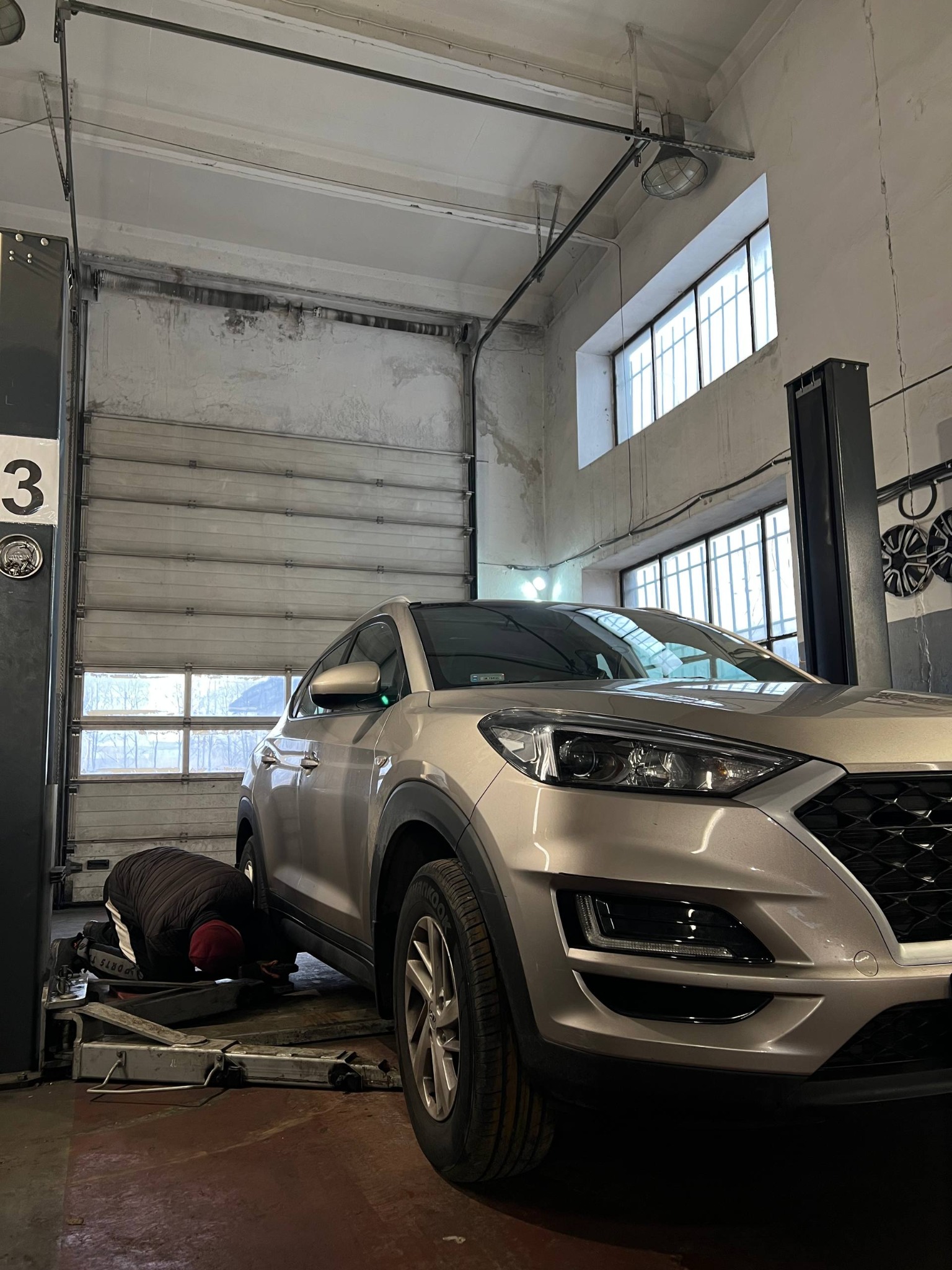 Mechanik w warsztacie samochodowym, pochylony pod srebrnym SUV-em na podnośniku, z otwartą bramą garażową w tle i oknami w górnej części ściany.