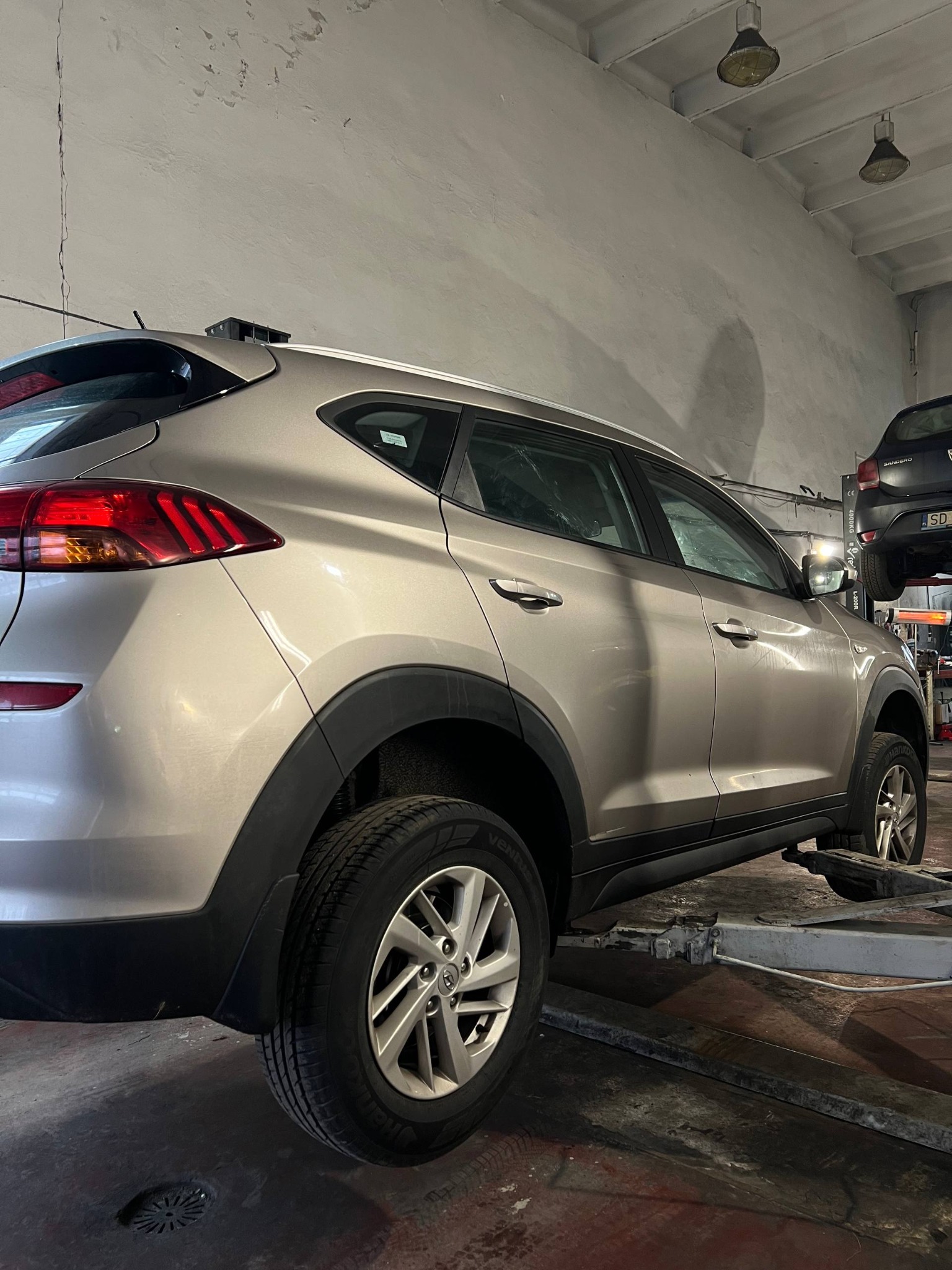 Srebrny Hyundai Tucson na podnośniku w warsztacie. Widoczny fragment drugiego auta na podnośniku w tle. Oświetlenie warsztatowe.
