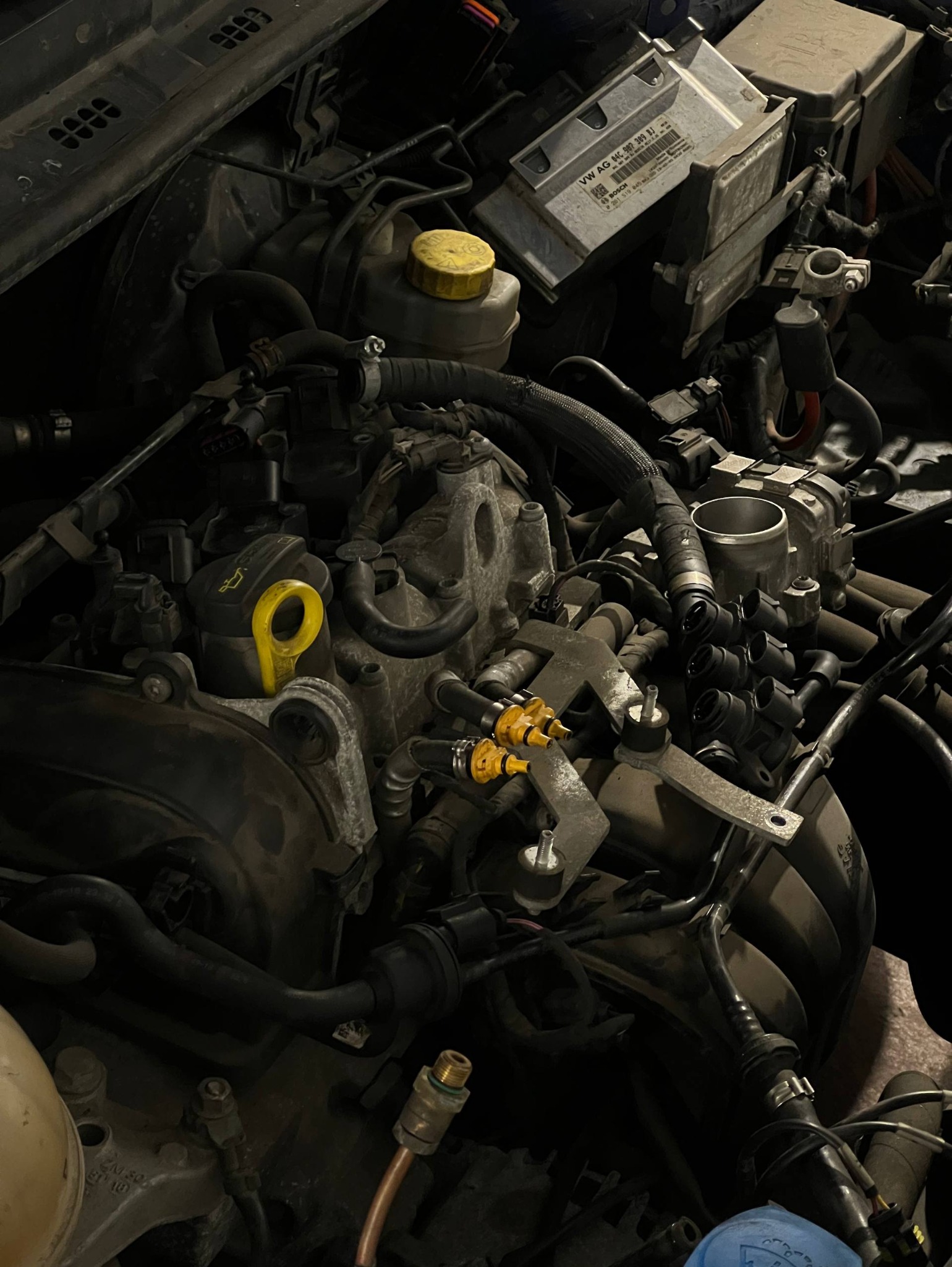 Otwarta maska samochodu z widocznym silnikiem i elementami układu chłodzenia. Żółte detale na tle ciemnych części. Widoczne przewody, złącza i zbiorniczki płynów.