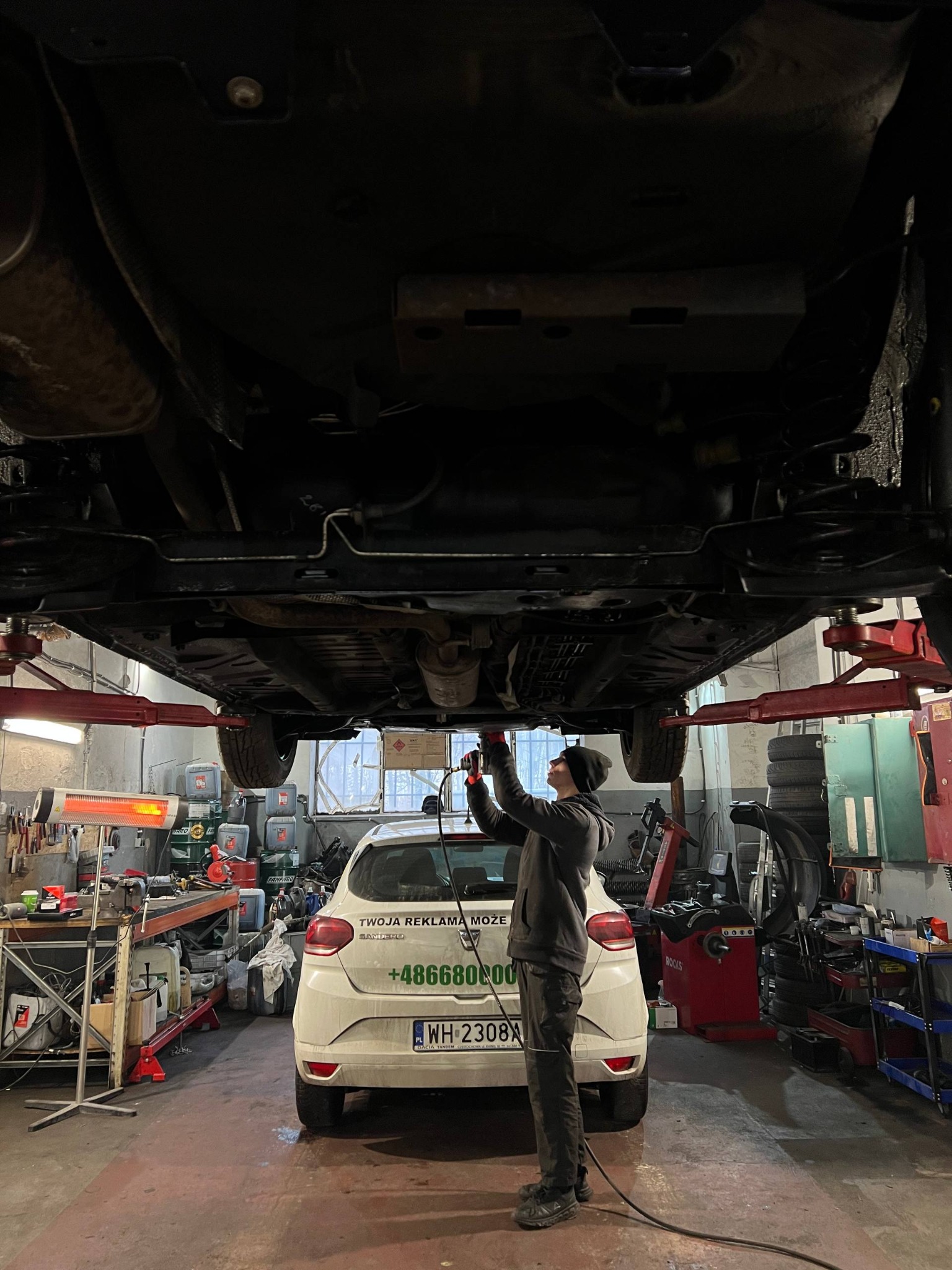 Mechanik w warsztacie samochodowym, w trakcie naprawy podwozia auta na podnośniku. Widoczny biały samochód z reklamą na tylnej klapie i narzędzia warsztatowe.