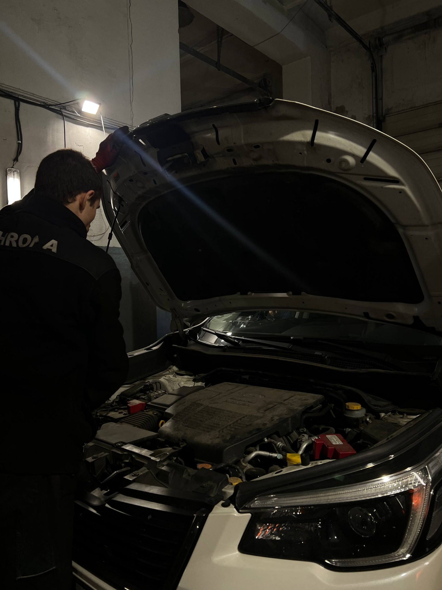 Mechanik w ciemnym warsztacie pochyla się nad otwartą maską białego samochodu, oświetlając silnik lampą warsztatową. Widoczny fragment logotypu na ubraniu.