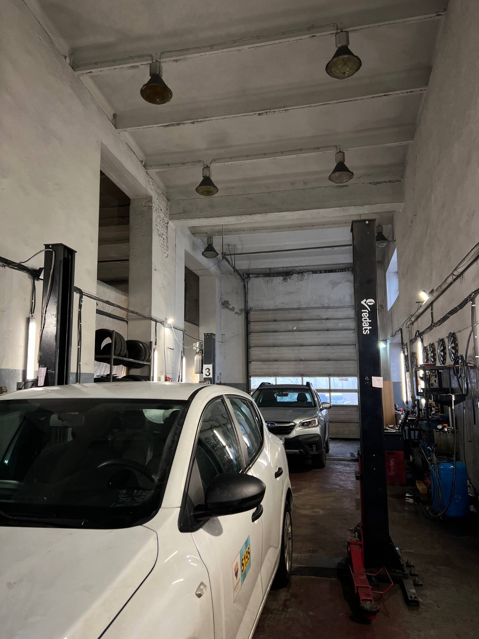 Wnętrze warsztatu samochodowego z dwoma autami: białym na pierwszym planie i srebrnym w tle, podnośnik kolumnowy, oświetlenie przemysłowe i opony na półce.