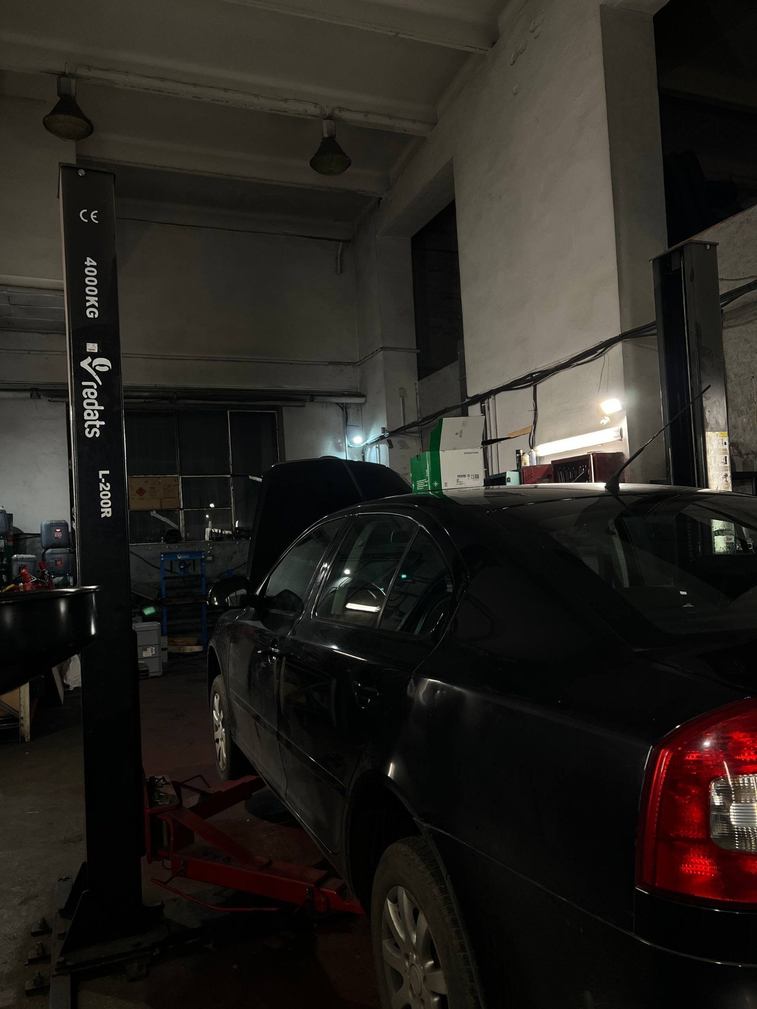 Czarny samochód na podnośniku w warsztacie. Otwarta maska, widoczne narzędzia i wyposażenie warsztatowe. Ujęcie z boku, oświetlenie warsztatowe.