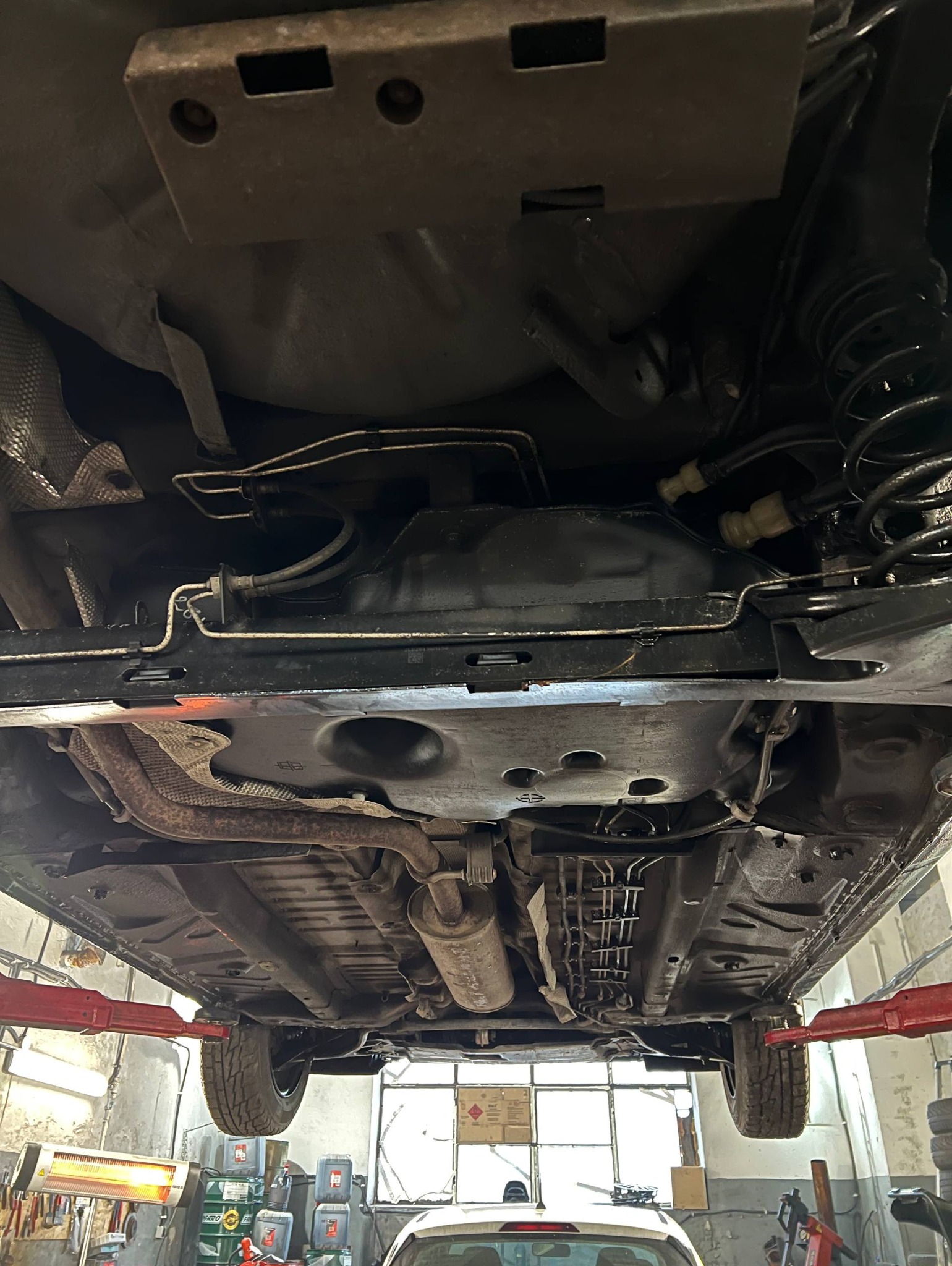 Podwozie samochodu na podnośniku w warsztacie, widok od spodu. Widoczne elementy zawieszenia, układu wydechowego i paliwowego. Oświetlenie warsztatowe.