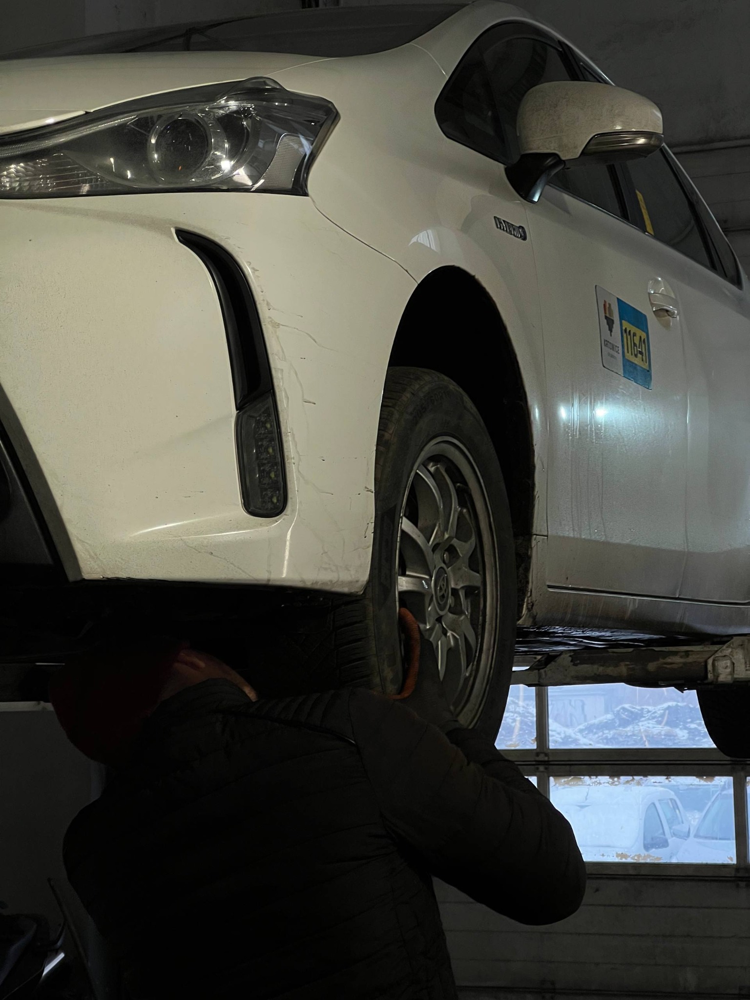 Biały samochód na podnośniku w warsztacie. Mechanik sprawdza koło. Widoczne okno warsztatu i inne pojazdy. Kontrola geometrii kół w serwisie.