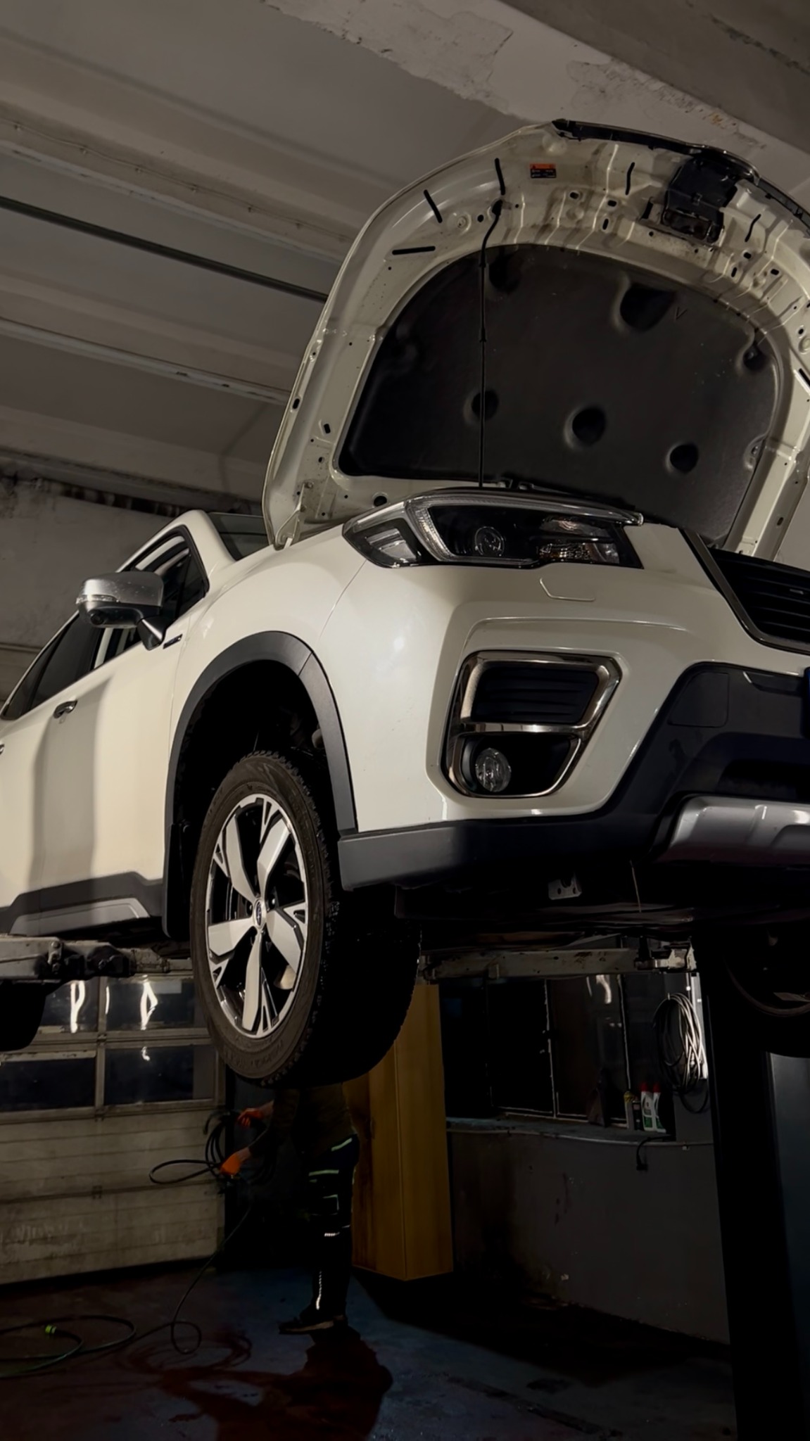 Biały samochód na podnośniku w warsztacie, z otwartą maską. Widoczne koło, oświetlenie warsztatowe i sylwetka mechanika. Ujęcie z dołu, podkreślające wysokość podnośnika.