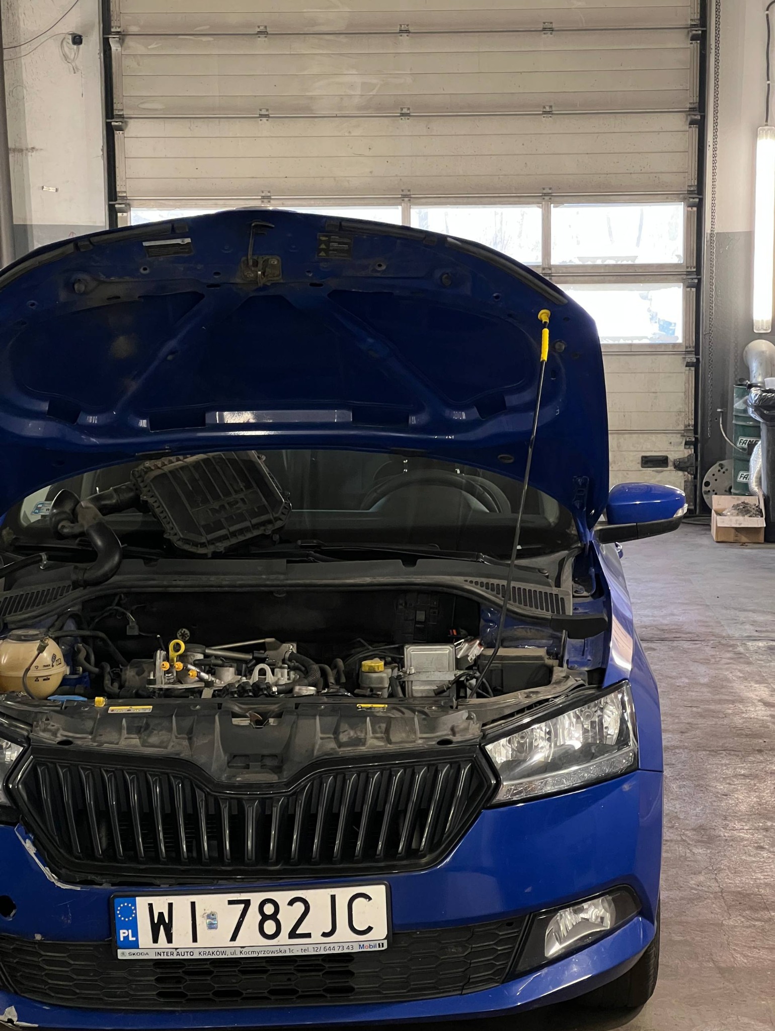 Niebieska Skoda z otwartą maską w warsztacie. Widoczny silnik i elementy mechaniczne. Żółty wskaźnik poziomu oleju. Tablica rejestracyjna z przodu. Brama garażowa w tle.