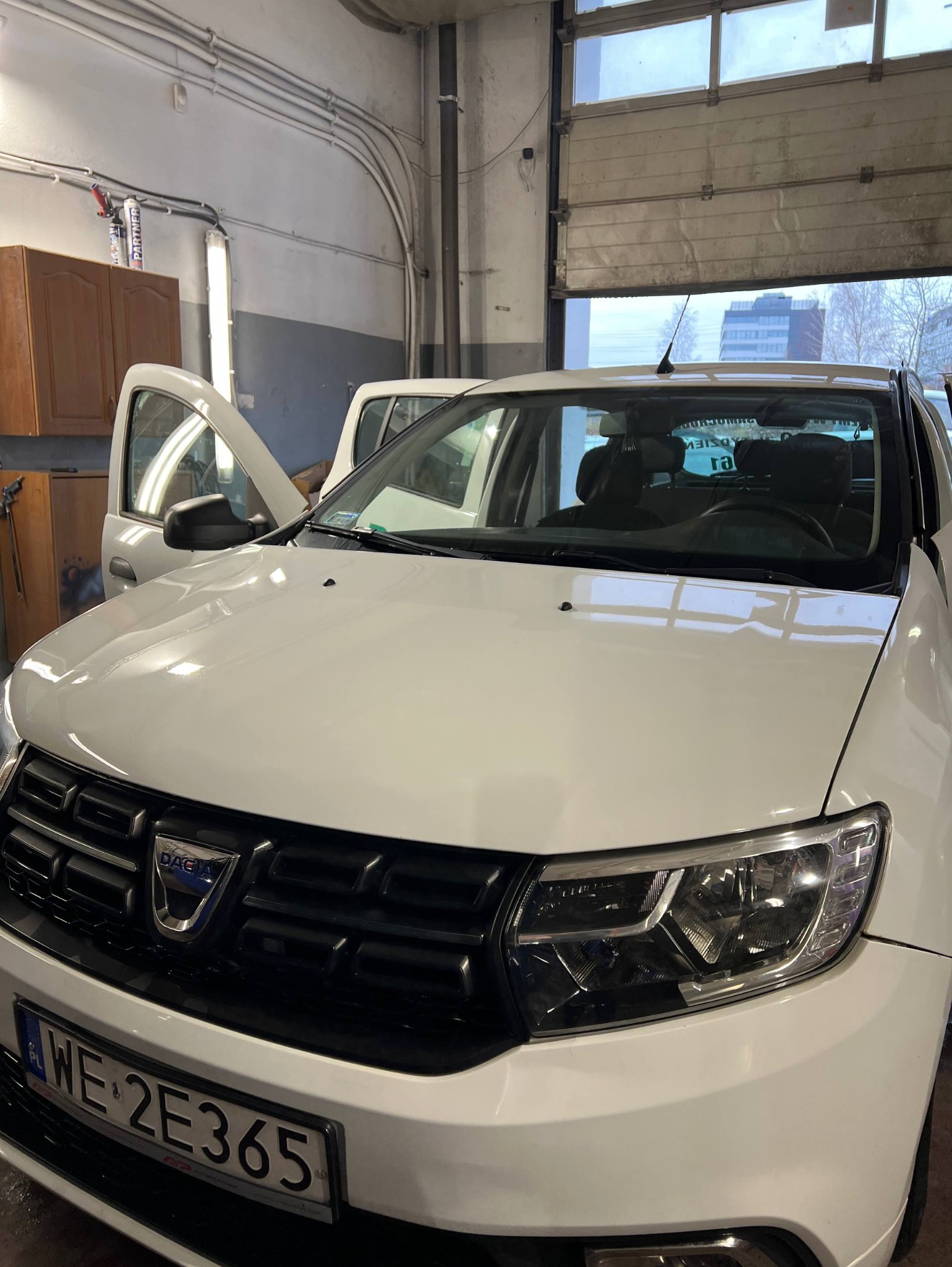 Biała Dacia Logan w warsztacie z otwartymi drzwiami, widoczny numer rejestracyjny WE 2E365, brama garażowa w tle. Diagnostyka pojazdu.
