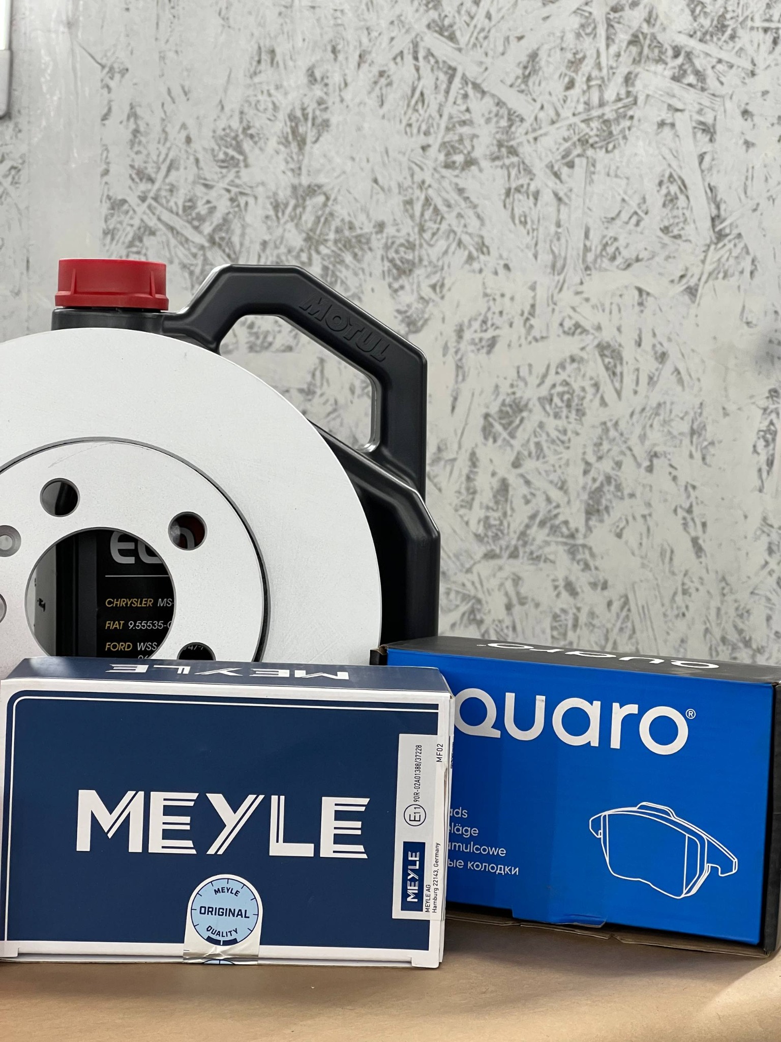 Nowe tarcze hamulcowe Meyle, klocki Quaro i olej Motul na tle ściany z płyty OSB. Zestaw części zamiennych do układu hamulcowego samochodu.
