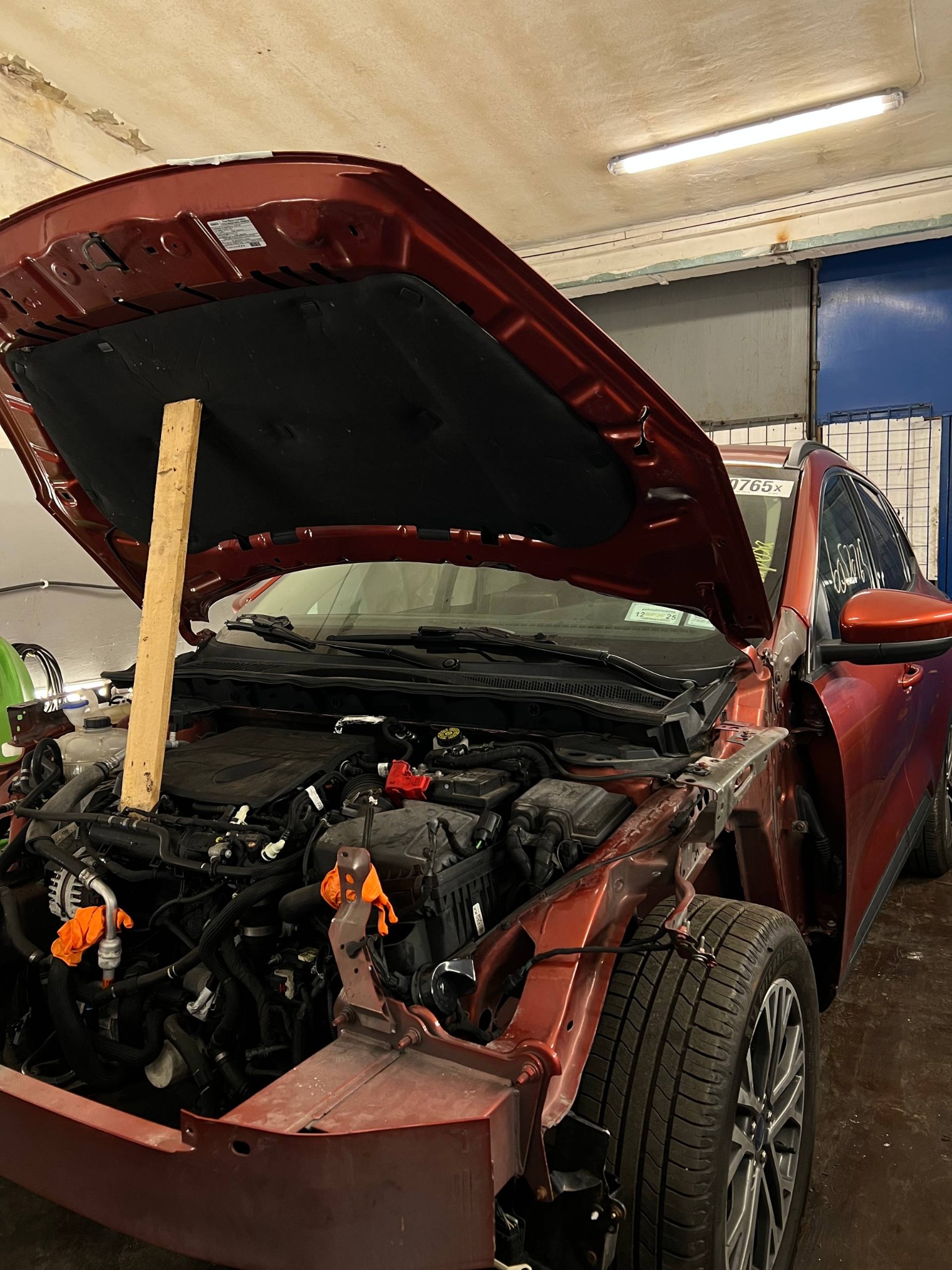 Czerwony samochód z otwartą maską, podpartą drewnianą deską. Widoczny silnik i elementy karoserii w warsztacie. Pomarańczowe rękawice na przewodach.