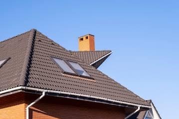 Ciemnobrązowy dach z dachówką karpiówką, okna dachowe, ceglany komin i rynna, na tle błękitnego nieba. Ujęcie z dołu, podkreślające solidność konstrukcji.