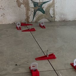 Hołyst - Układanie płytek podłogowych z użyciem systemu poziomowania, widoczne czerwone kliny i białe klipsy. W tle fragment ściany z dekoracyjnymi, wzorzystymi płytkami.