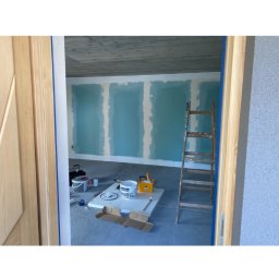 Hołyst - Wnętrze w trakcie prac: ściany z płyt gipsowo-kartonowych pomalowane na niebiesko, drabina oparta o ścianę, widoczne narzędzia i materiały malarskie. Surowy betonowy sufit.