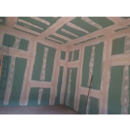 Hołyst - Ściany i sufit w trakcie przygotowania do malowania, pokryte zieloną farbą i białymi pasami szpachli, widoczne elementy konstrukcyjne. W tle drabina.