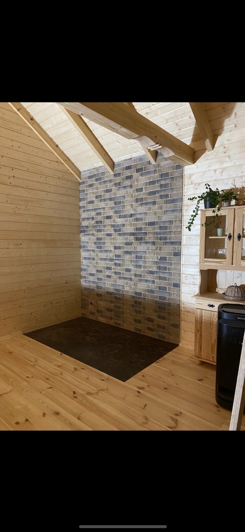 Drewniane wnętrze z akcentem ceglanej ściany i ciemną podłogą. Ustawiona szafka z roślinami. Skośny sufit z widocznymi belkami. Nietypowe wykończenie.