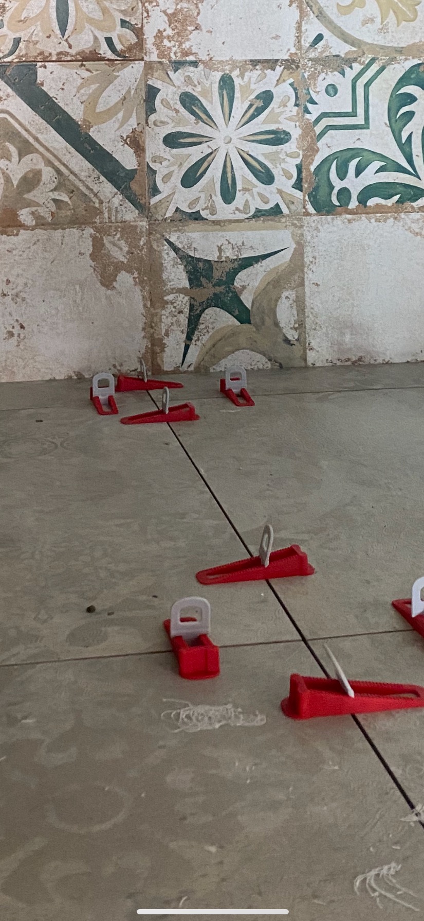 Układanie płytek podłogowych z użyciem systemu poziomowania, widoczne czerwone kliny i białe klipsy. W tle fragment ściany z dekoracyjnymi, wzorzystymi płytkami.