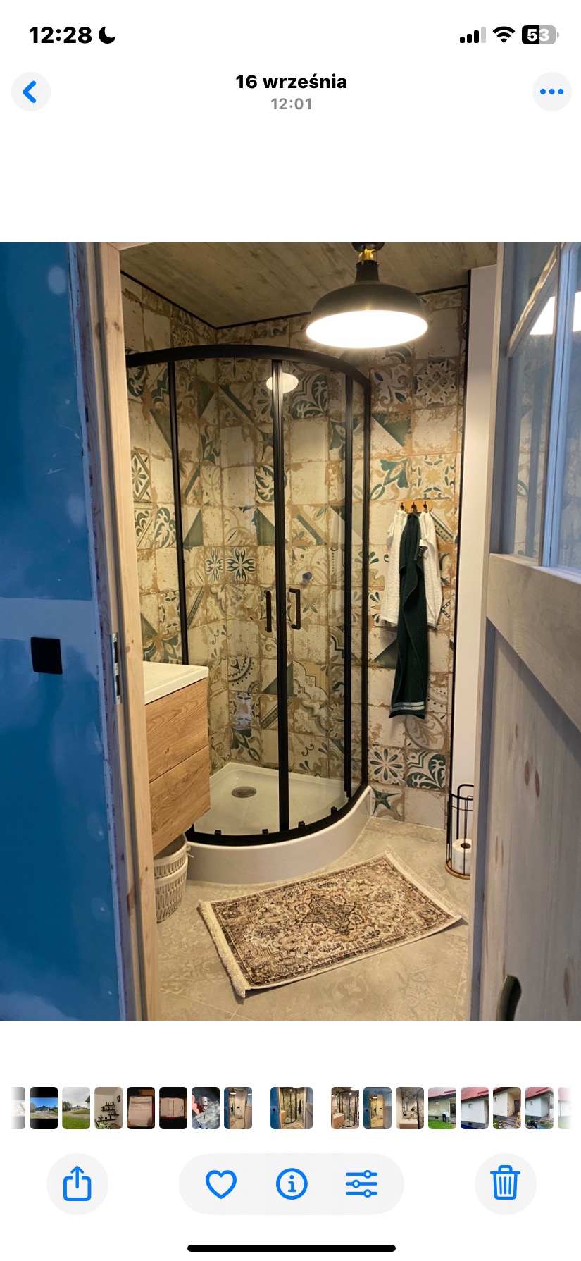 Wykończona łazienka z kabiną prysznicową w czarnej ramie, wzorzystymi płytkami i drewnianą szafką pod umywalką. Na podłodze leży dywanik.