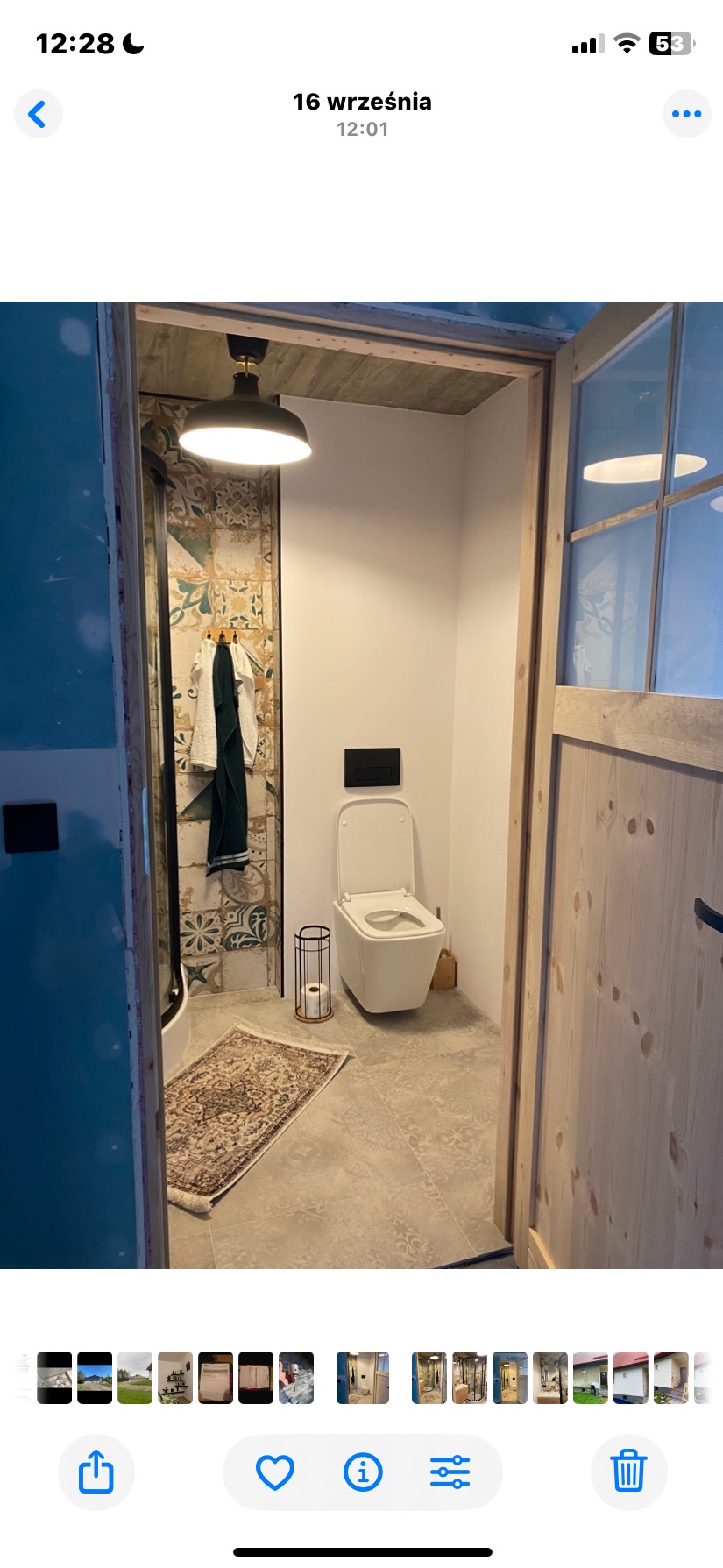 Wykończona łazienka z kabiną prysznicową z mozaiką, wiszącą toaletą i drewnianymi drzwiami. Widok z zewnątrz, z dywanikiem na podłodze.