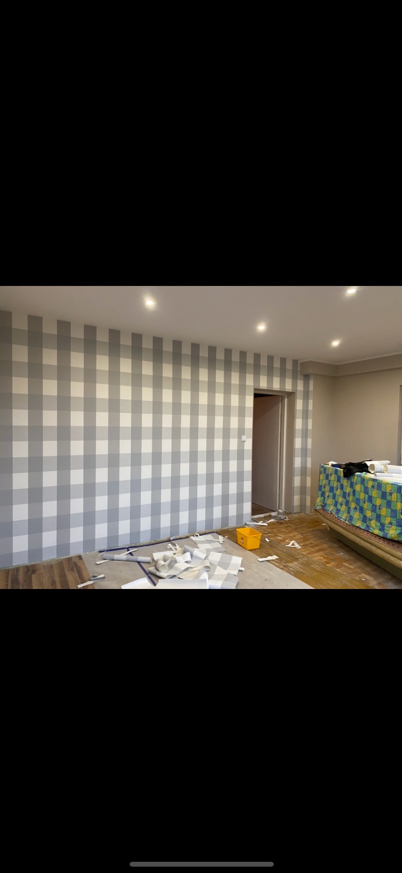 Pokój po tapetowaniu w szaro-białą kratę. Na podłodze resztki tapety i żółte wiadro. Widoczne oświetlenie sufitowe i fragment sąsiedniego pomieszczenia.