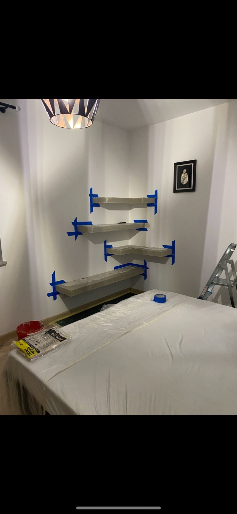 Montaż półek narożnych w pokoju, zabezpieczonym folią malarską. Półki przymocowane taśmą malarską, widoczna drabina i lampa sufitowa. Etap prac wykończeniowych.