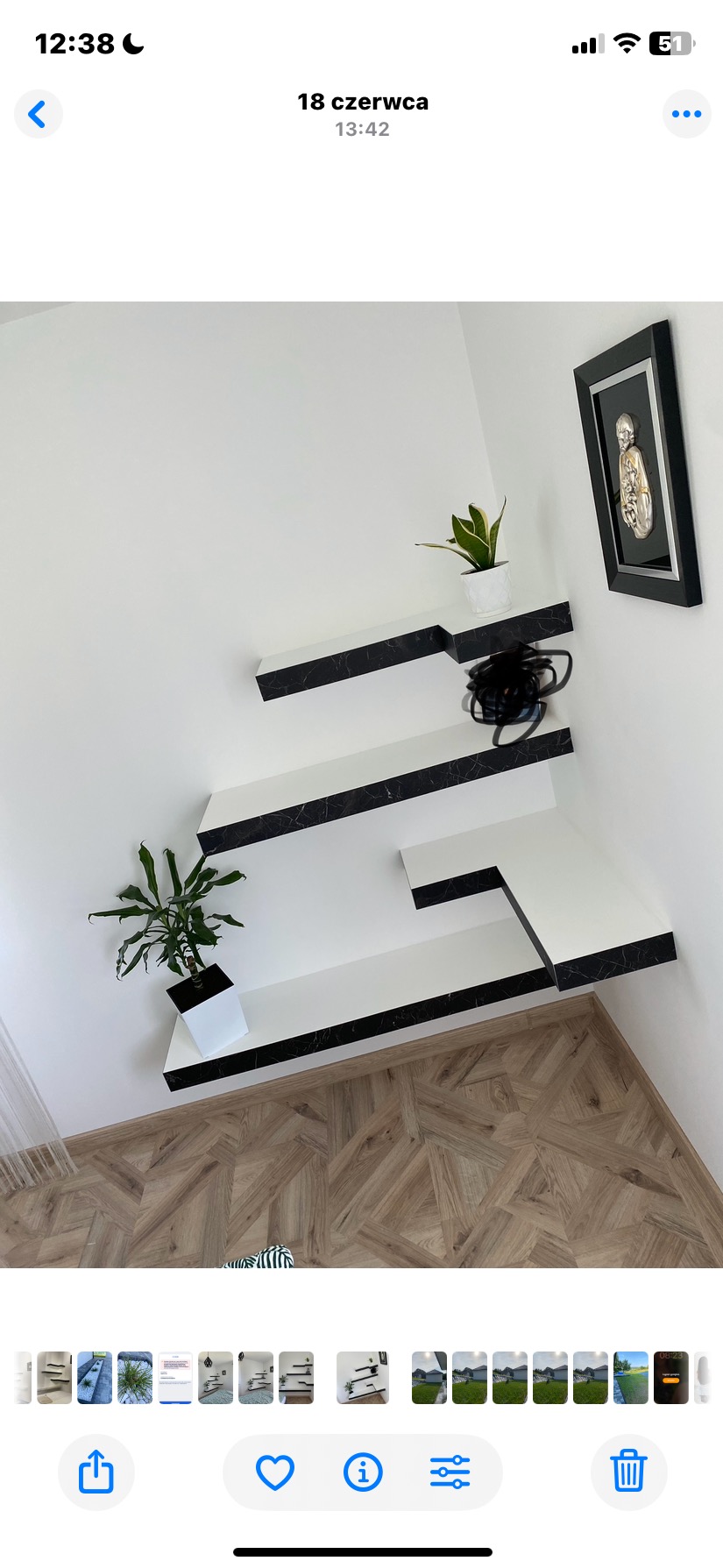 Białe, asymetryczne półki z czarnym marmurkowym wzorem na ścianie, z roślinami doniczkowymi i obrazem w czarnej ramie. Parkiet w jodełkę na podłodze.