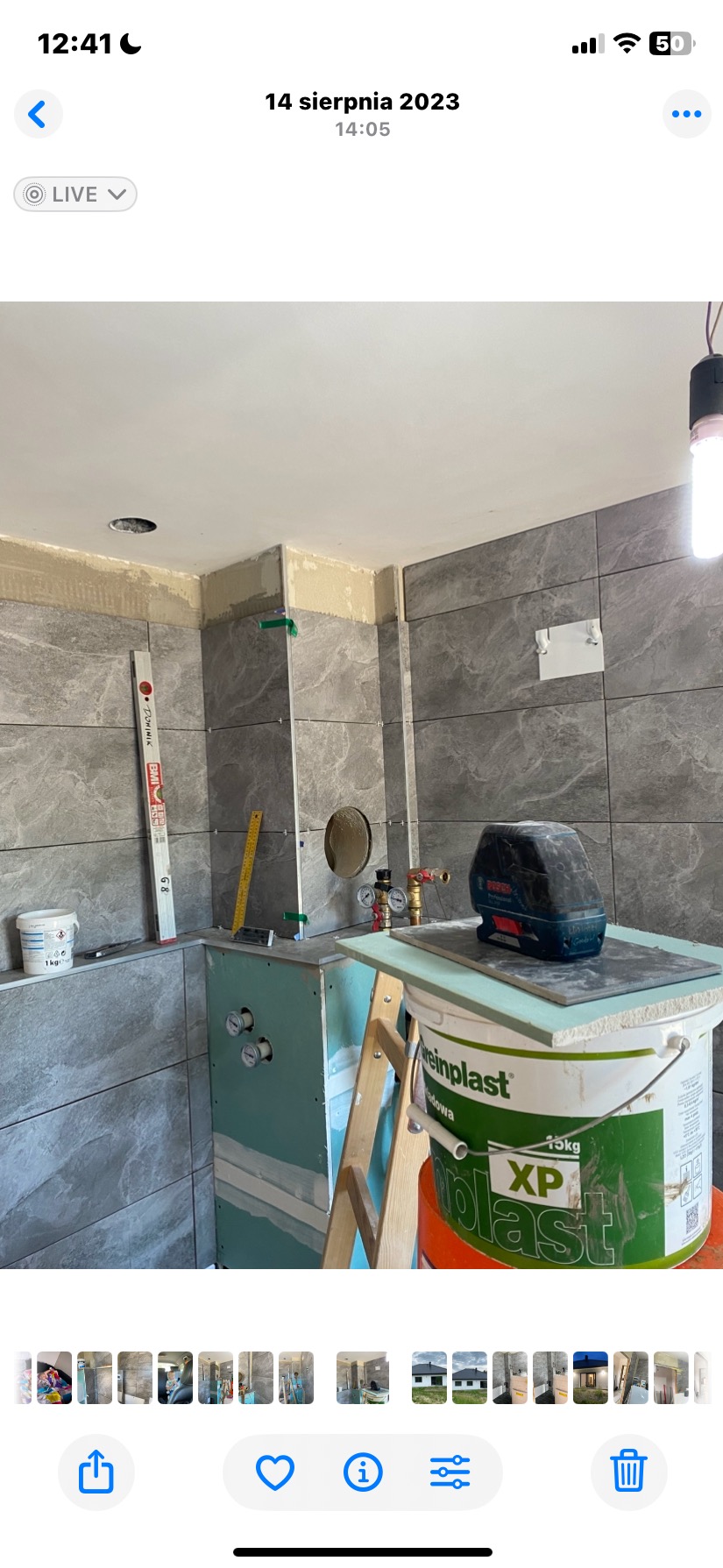 Łazienka w trakcie remontu: szare płytki imitujące kamień na ścianach, zabudowa z płyt gips-karton, poziomica, wiadro z zaprawą i laser krzyżowy na drabinie.