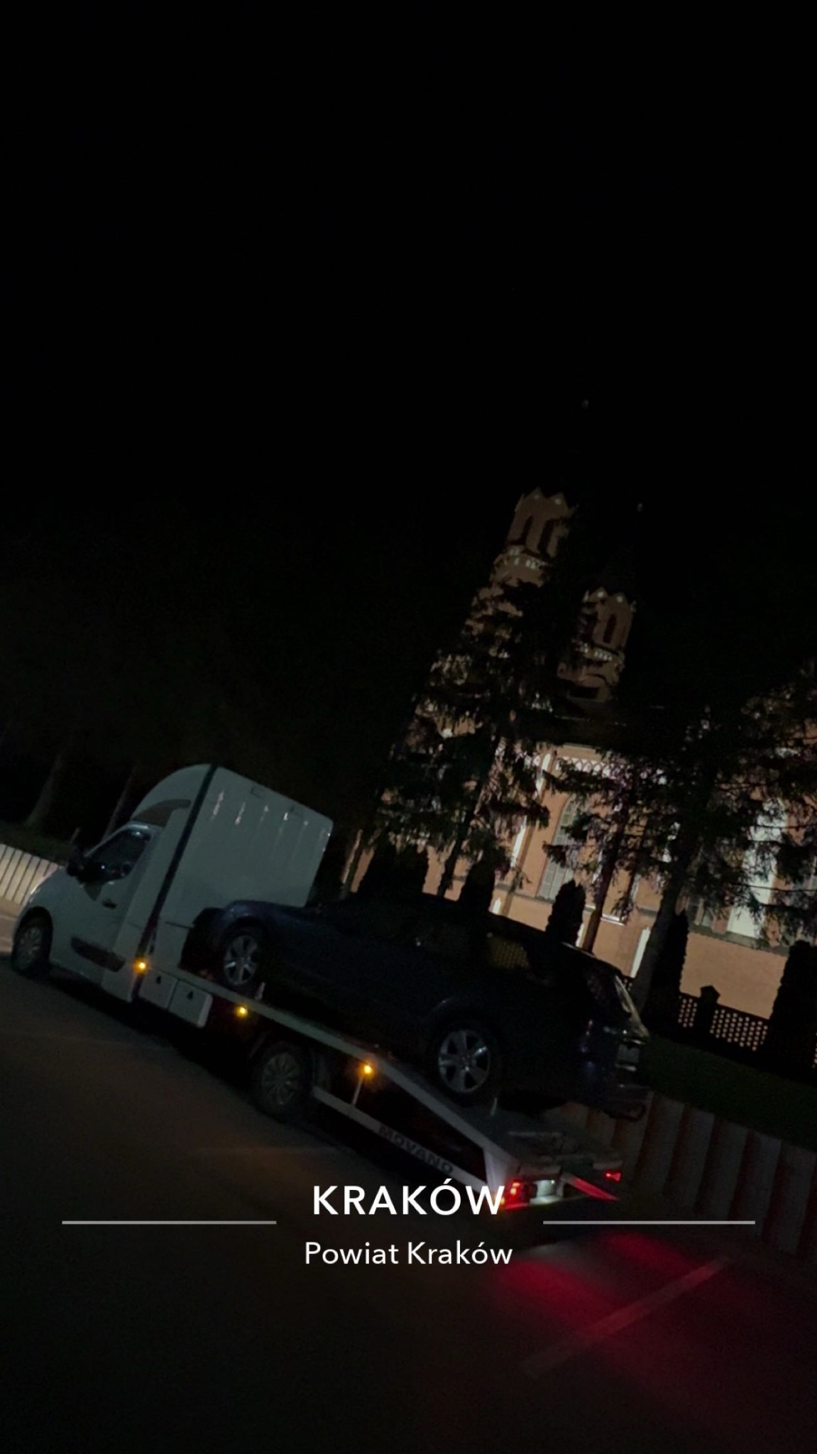 Nocny transport uszkodzonego auta na lawecie w Krakowie. W tle widoczny budynek z wieżą. Ujęcie z dołu, podkreślające miejski krajobraz i specyfikę usługi.