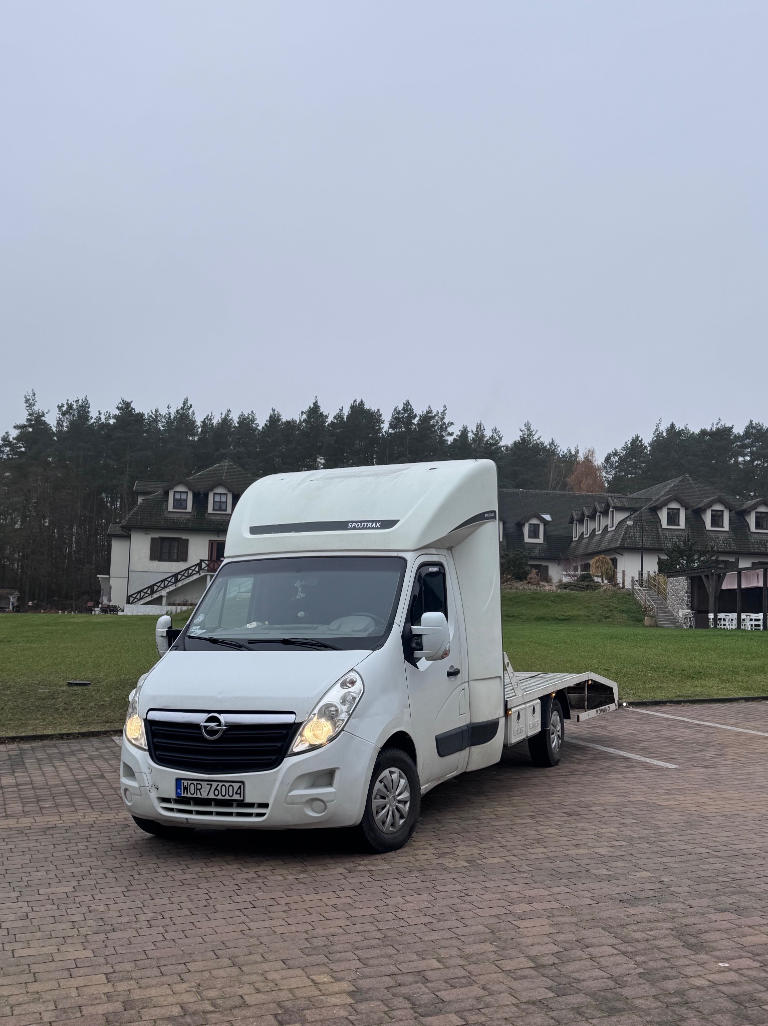 Biała laweta Opel Movano na parkingu z kostki, z włączonymi światłami, na tle budynku z zielonym trawnikiem i lasem w pochmurny dzień. Widok z przodu.