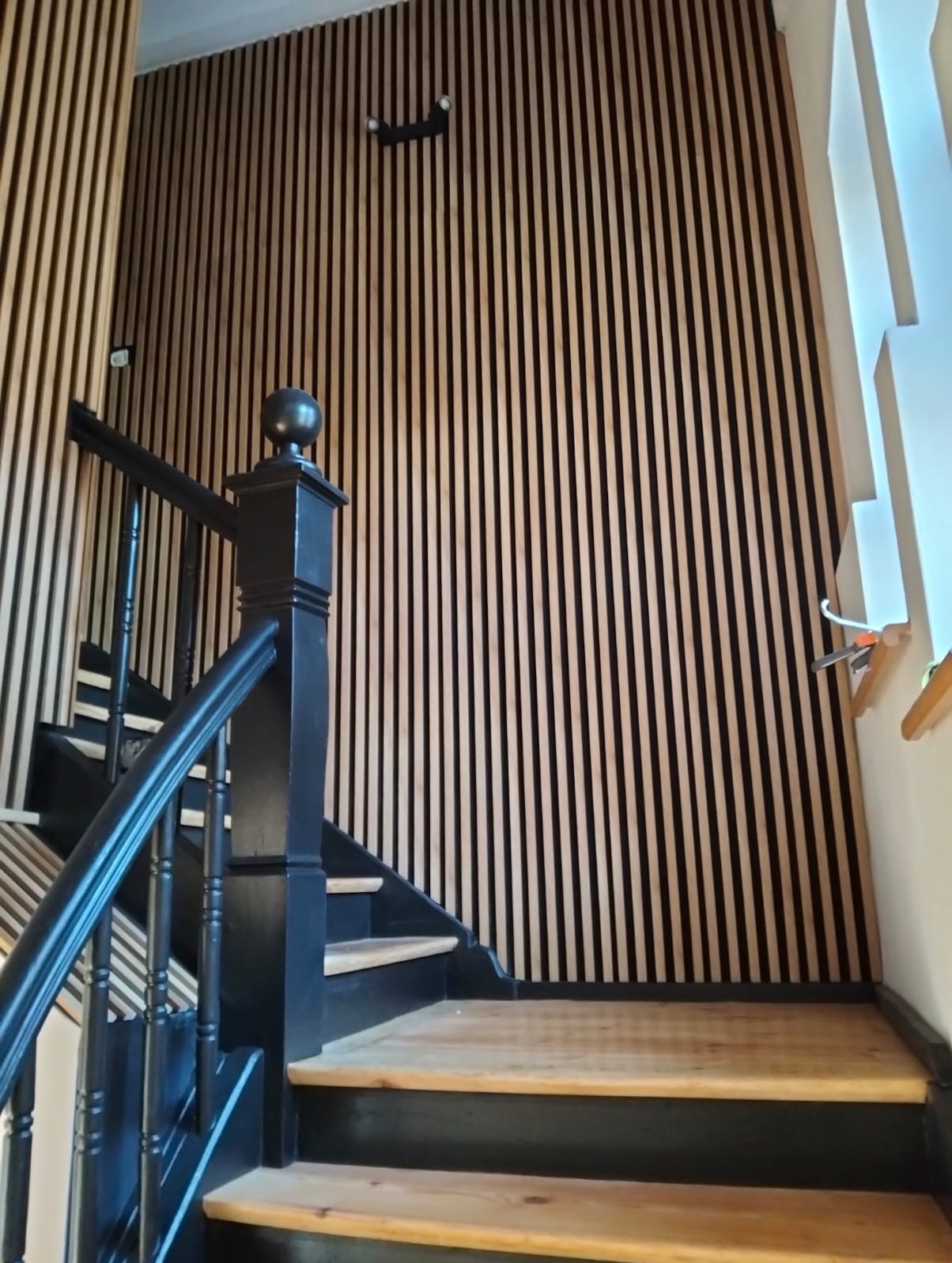 Schody z czarną balustradą i drewnianymi stopniami na tle ściany z dekoracyjnych paneli pionowych. Widoczne oświetlenie sufitowe.