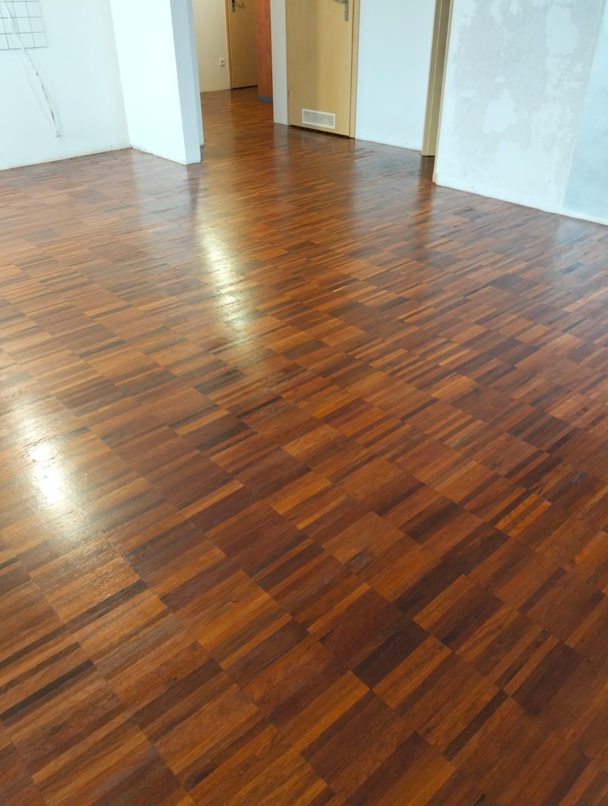 Błyszcząca, świeżo wycyklinowana podłoga parkietowa z ciemnego drewna ułożona we wzór szachownicy, widok z perspektywy podłogi, z widocznymi drzwiami w tle.