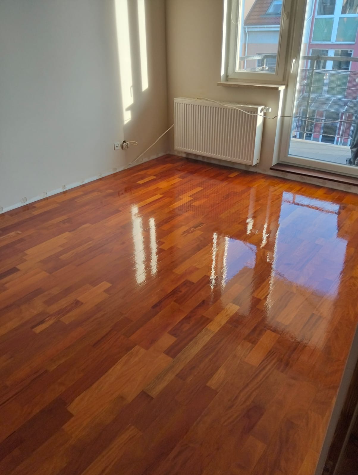Błyszczący, świeżo polakierowany parkiet w pokoju z grzejnikiem i oknem. Parkiet ułożony w klasyczny wzór, odbija światło okienne. Widoczne gniazdka elektryczne przy ścianie.