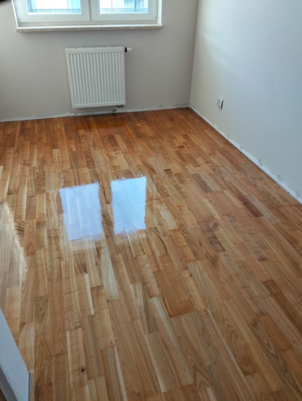 Błyszczący parkiet po cyklinowaniu w jasnym pokoju z grzejnikiem i oknem. Widoczne refleksy światła na gładkiej powierzchni drewna. Świeżo wykończona podłoga.